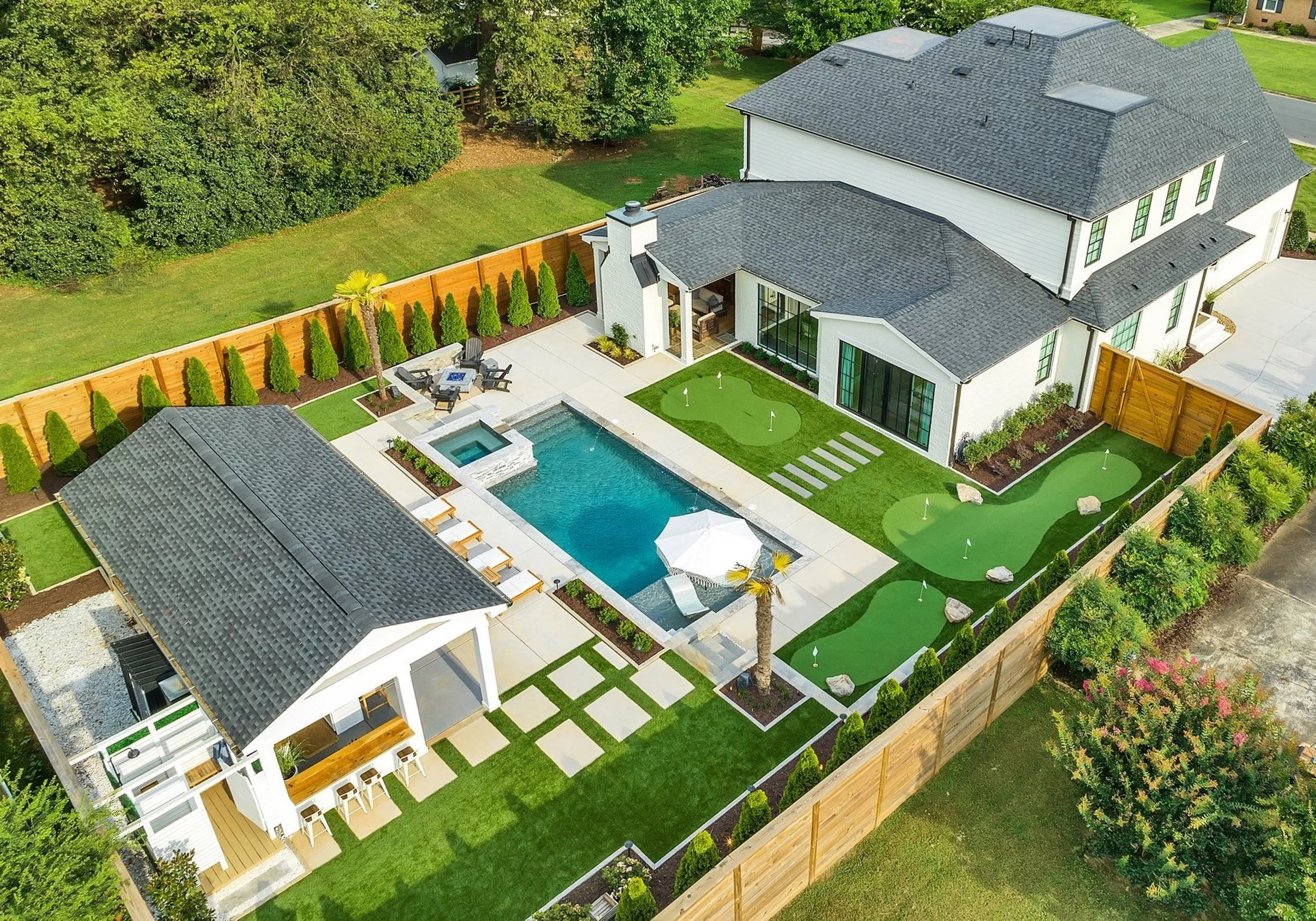 A backyard with a swimming pool, a small hot tub, and a putting green, enclosed by a wooden fence, with a house, a poolside cabana, and landscaped lawn and trees.