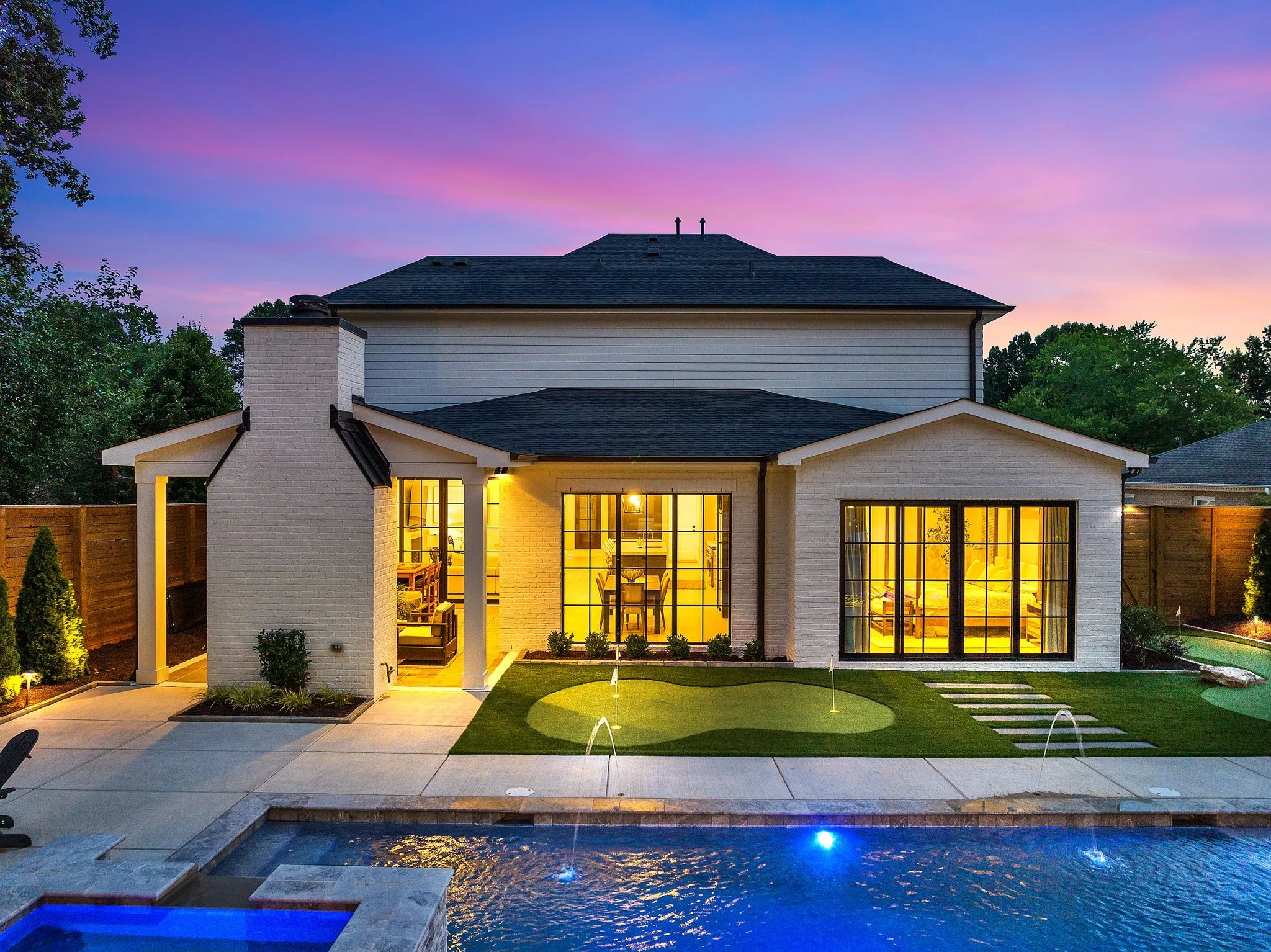 Backyard of a house with a swimming pool, outdoor seating, a mini putt-putt golf area, and illuminated interior, during dusk with a colorful sky.