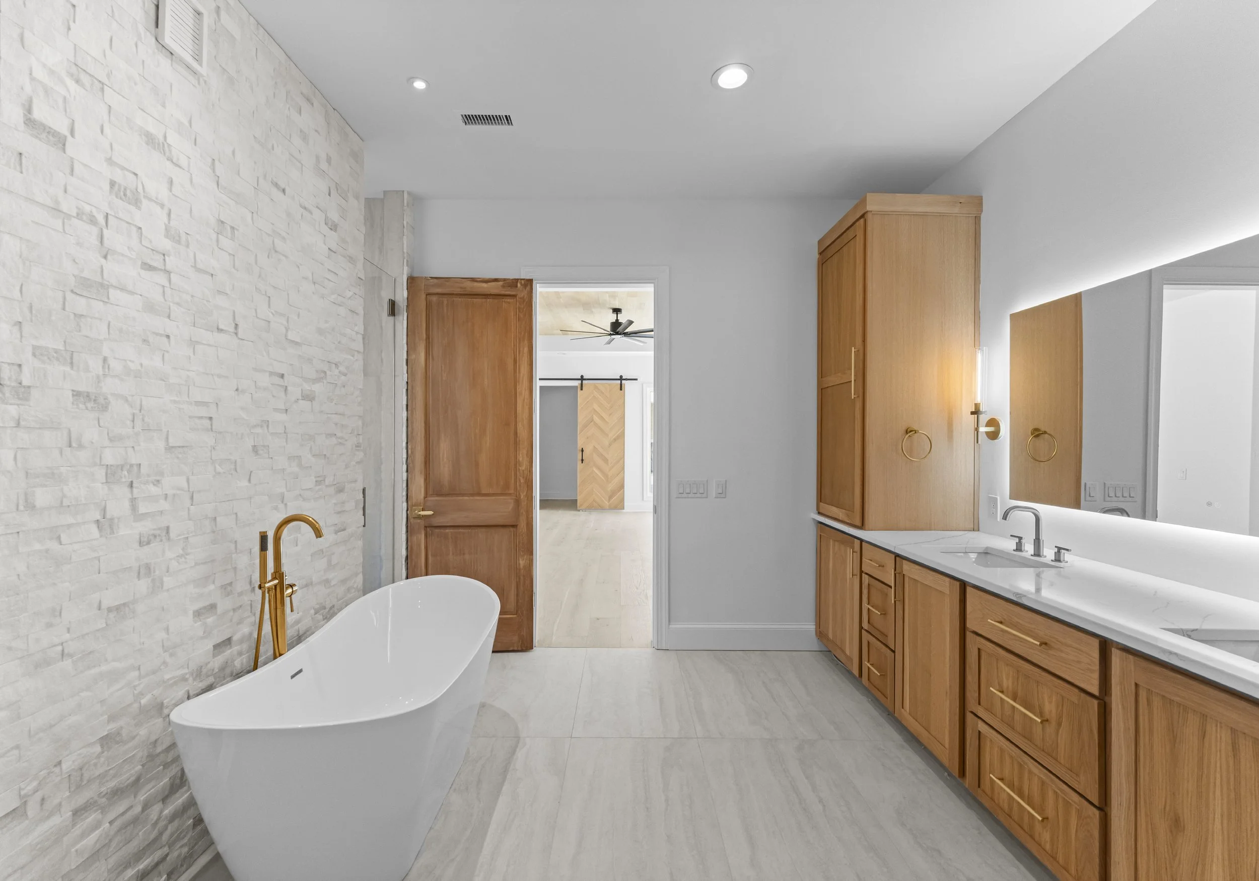 Modern bathroom with white freestanding bathtub, wooden cabinetry, marble countertop, large mirror, and a textured white brick wall, with door leading to another room.
