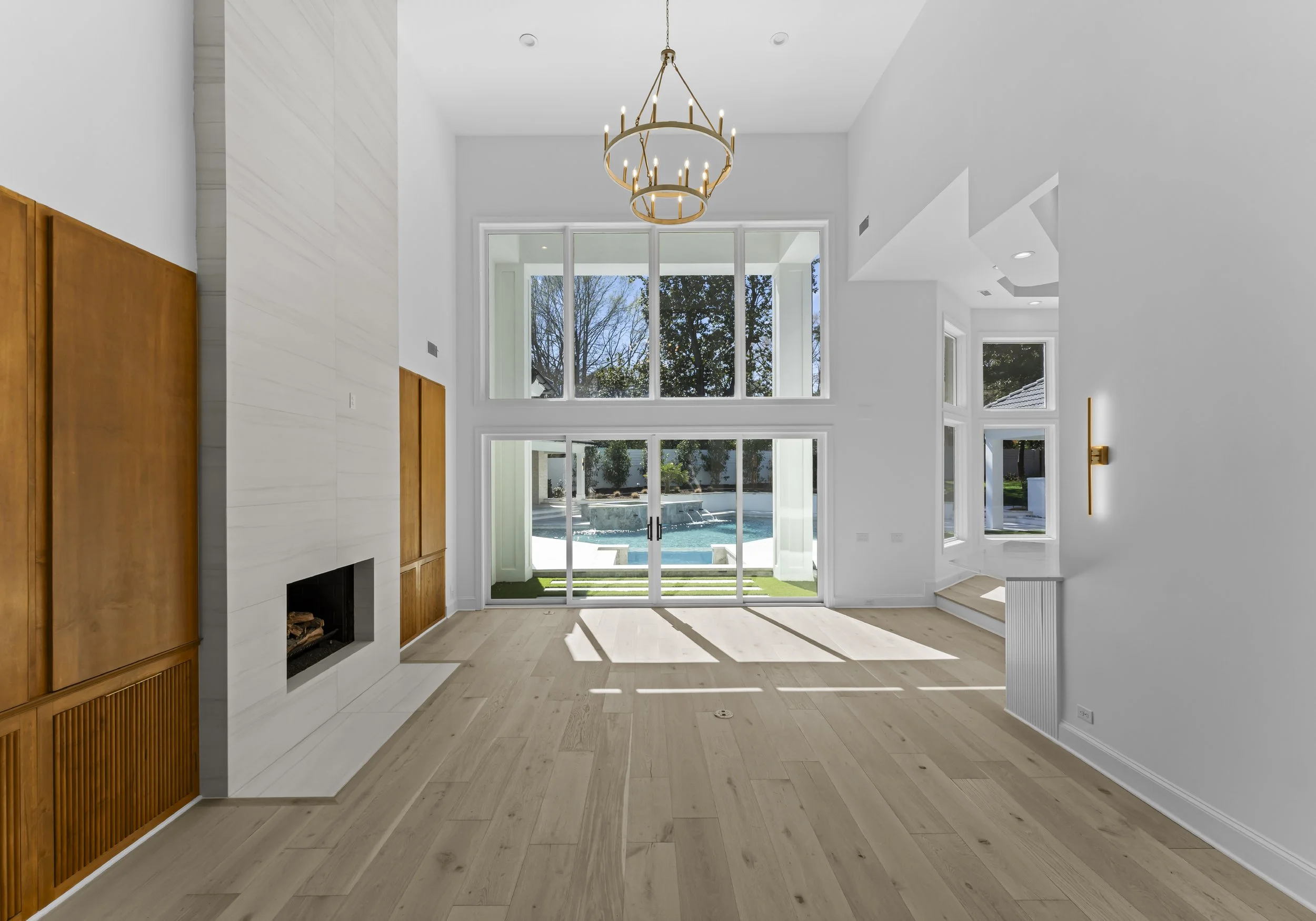 Empty living room with high ceiling, large windows overlooking a pool, and a modern chandelier hanging from the ceiling.