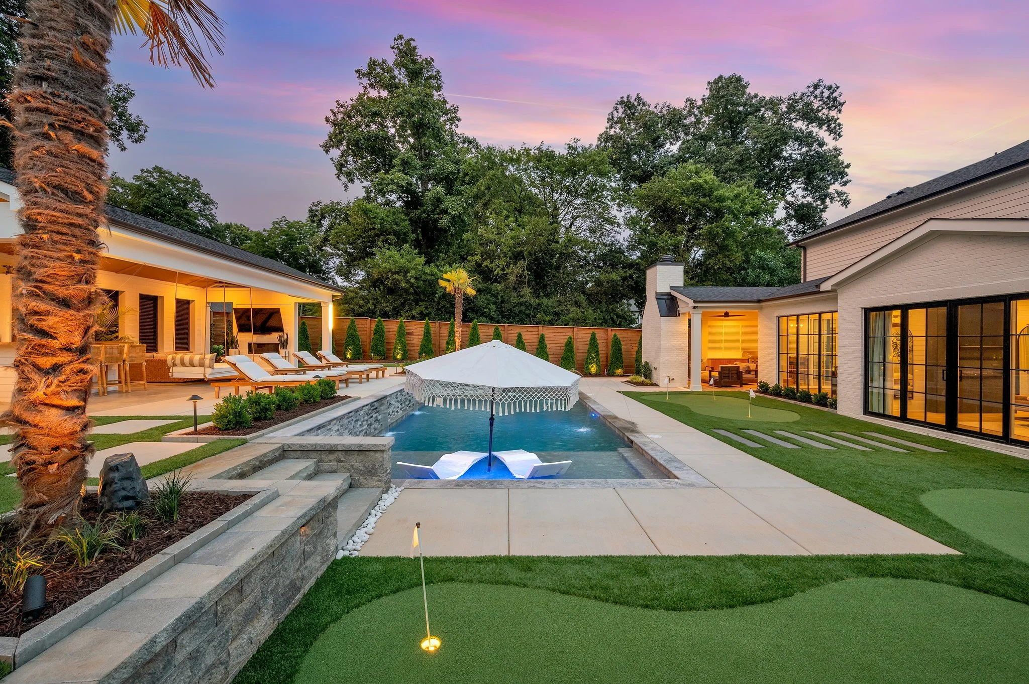 A backyard with a rectangular swimming pool, white lounge chairs, an umbrella, and a small putting green. The yard is surrounded by a wooden fence, with a house featuring large glass doors and a patio area illuminated with warm lighting. There are tr
