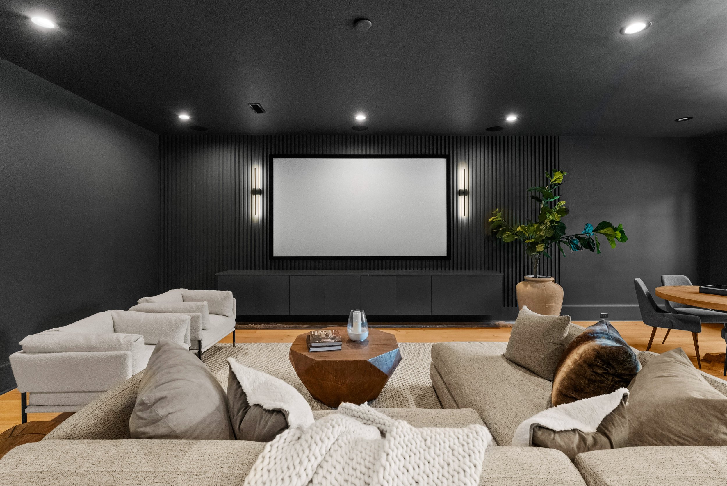 Modern living room with beige sofas, a geometric wooden coffee table, a large framed television on a black textured wall, a tall potted plant, and dining chairs in the background.
