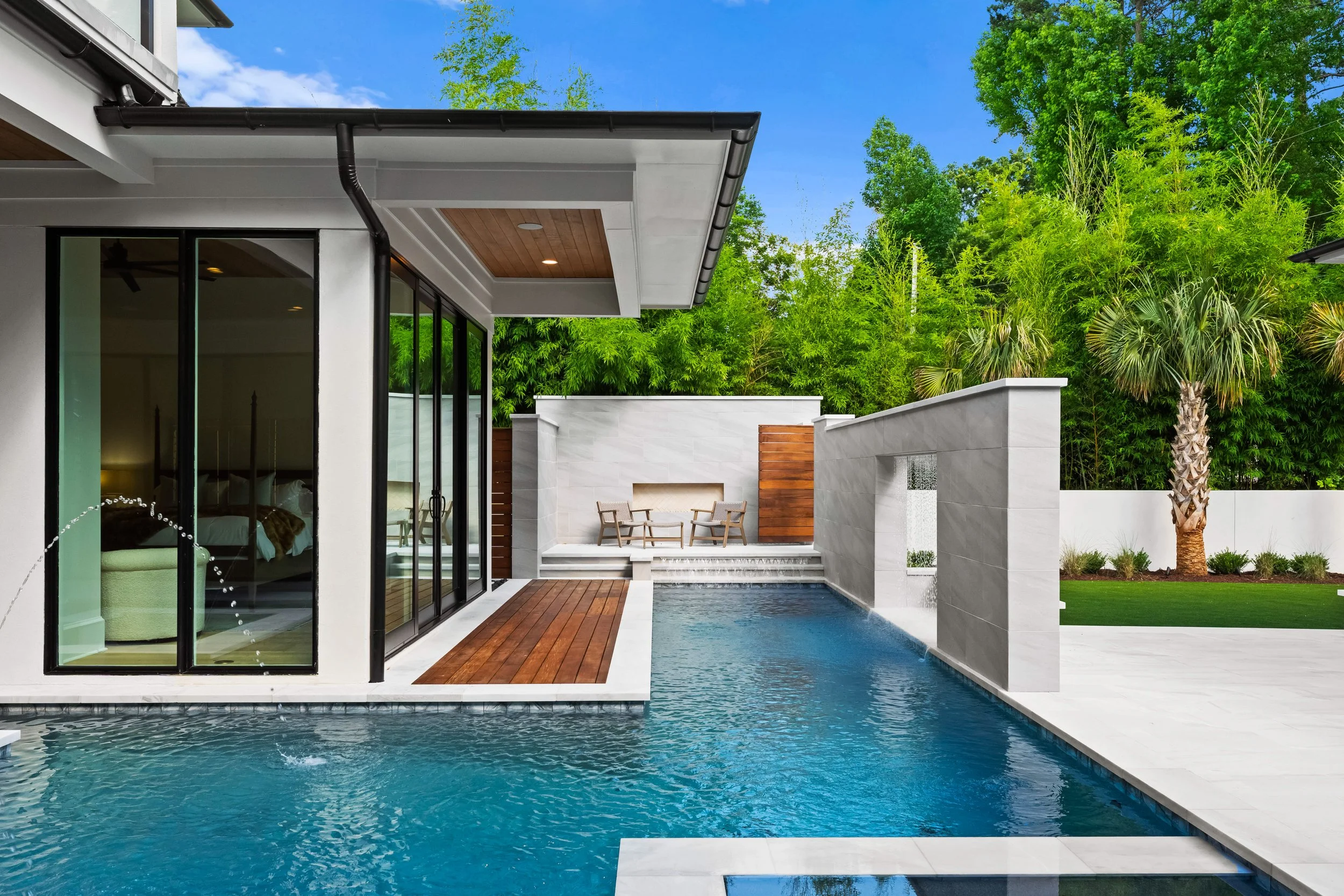 Modern backyard with a swimming pool, wooden deck, and outdoor seating area. House features large glass doors, white walls, and a water wall feature. Surrounding greenery includes trees and well-manicured grass.