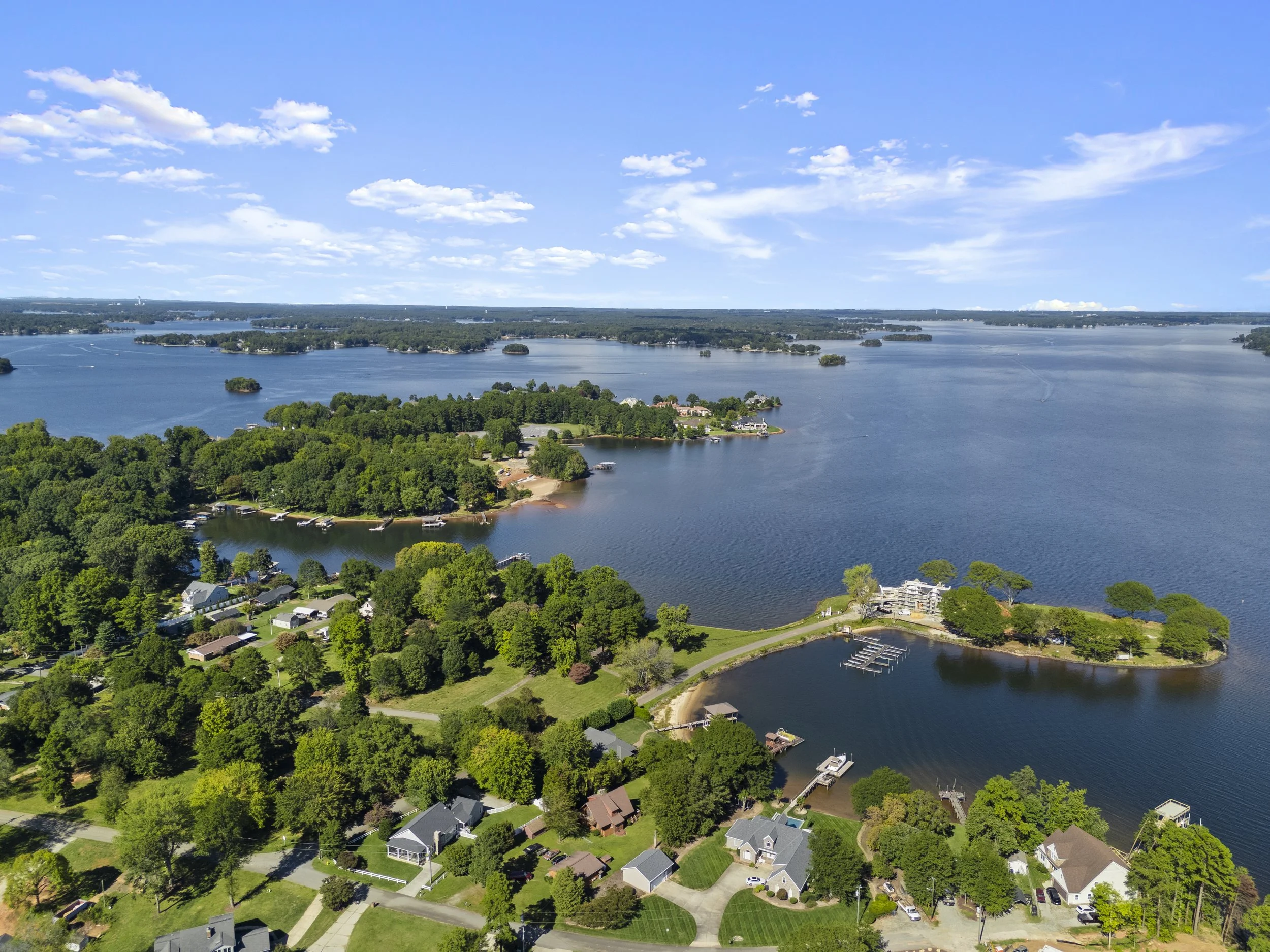Aerial view of a lakeside community with houses, trees, docks, and boats on a large body of water under a partly cloudy sky.