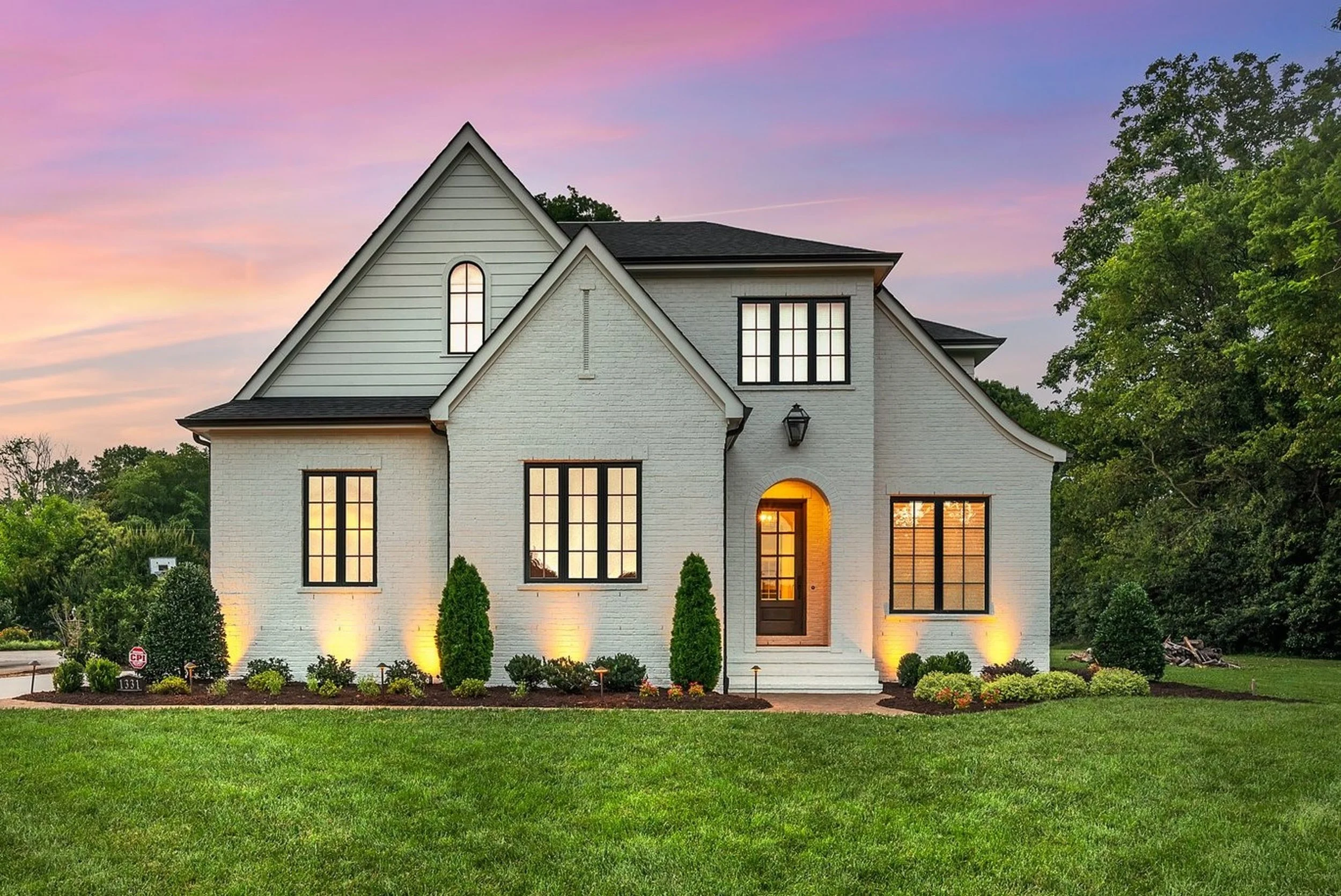 A modern white two-story house with large windows, a front porch, and landscaped front yard with bushes and grass at sunset.