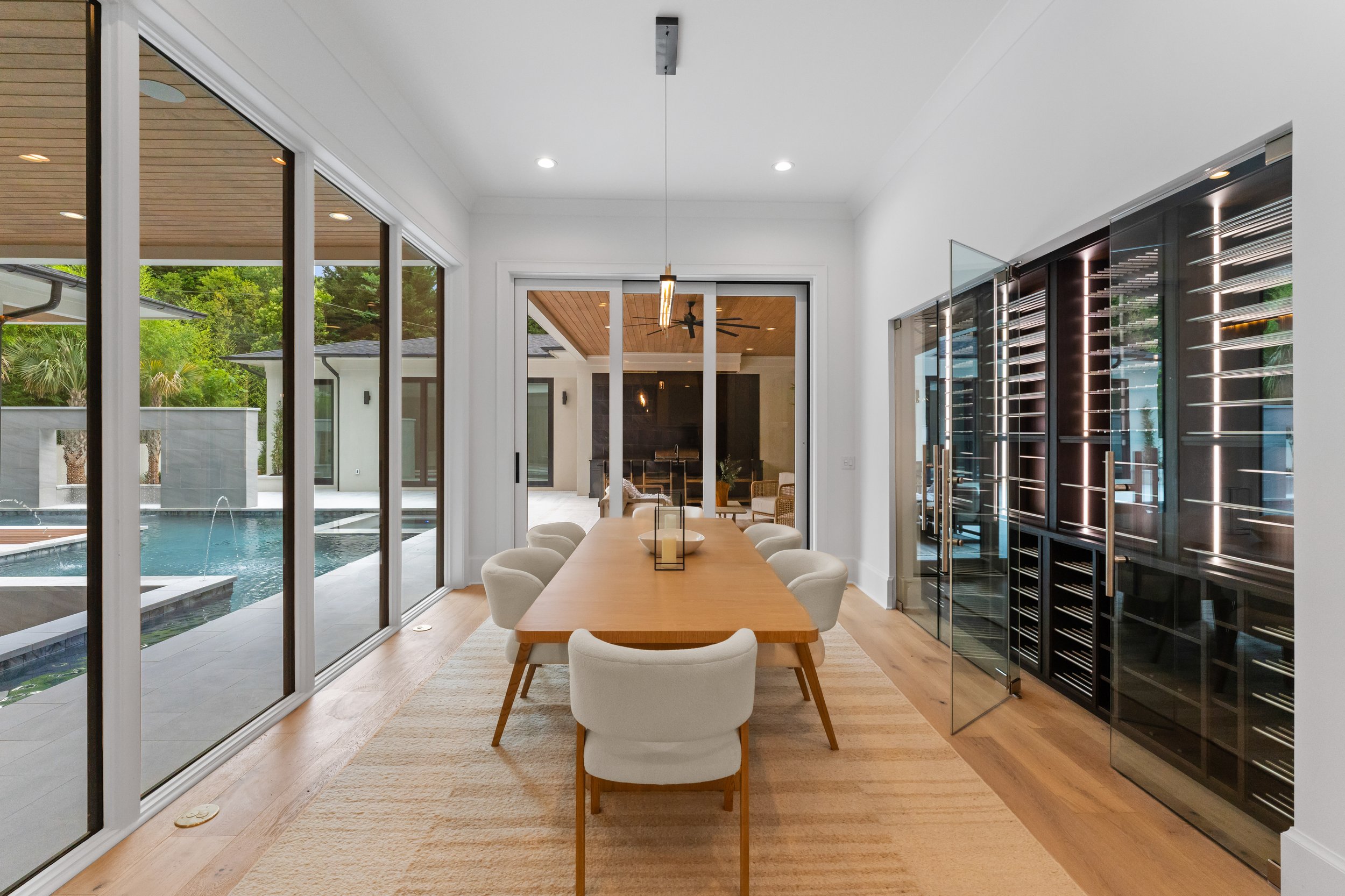 Modern dining room with large glass sliding doors overlooking a pool and yard, wooden dining table with white cushioned chairs, and wine storage cabinet with glass door on right.