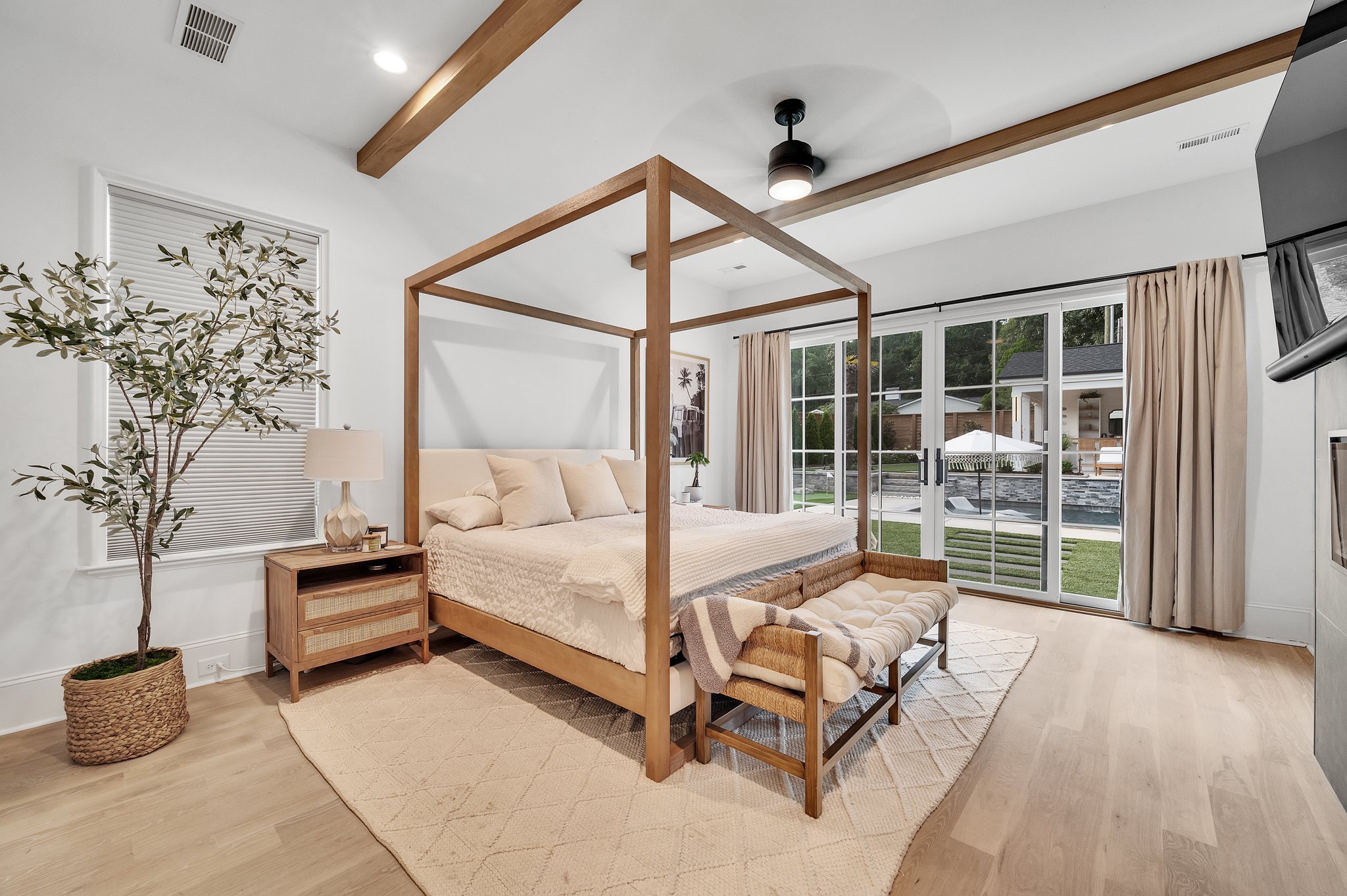 Modern bedroom with a wooden canopy bed, nightstand, and sliding glass doors leading to a backyard with a pool. Neutral tones, large window, and minimal decor.