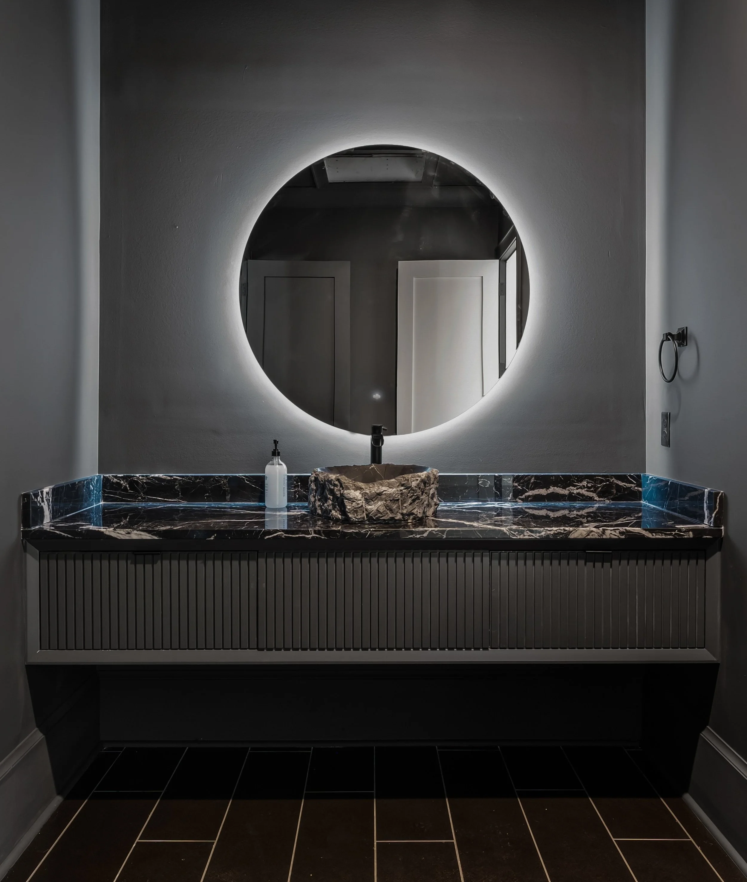Modern bathroom with a round illuminated mirror, black marble sink with a stone basin, white wall, black tiled floor, and soap dispenser.