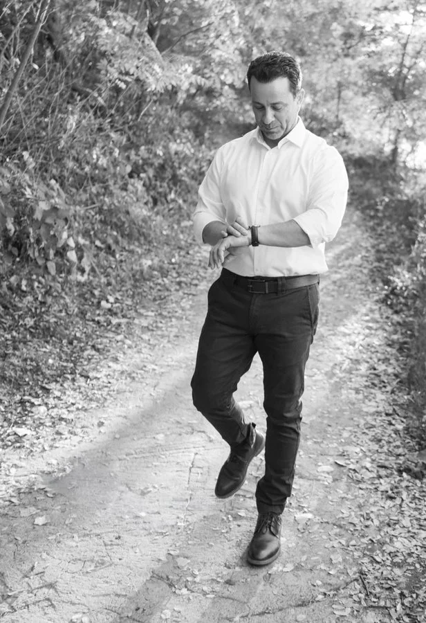 A man in a white shirt and dark pants walking on a dirt path in a wooded area, looking at his watch.