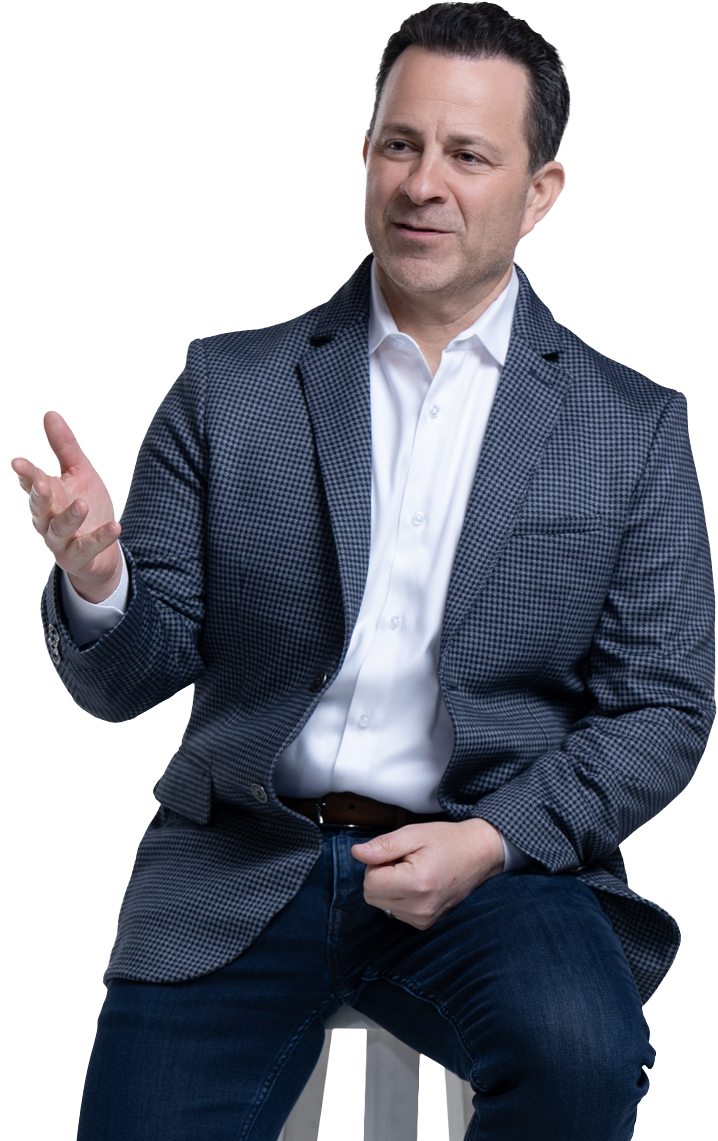A man wearing a checked blazer and white shirt, sitting and gesturing with his right hand, dark hair and fair skin.