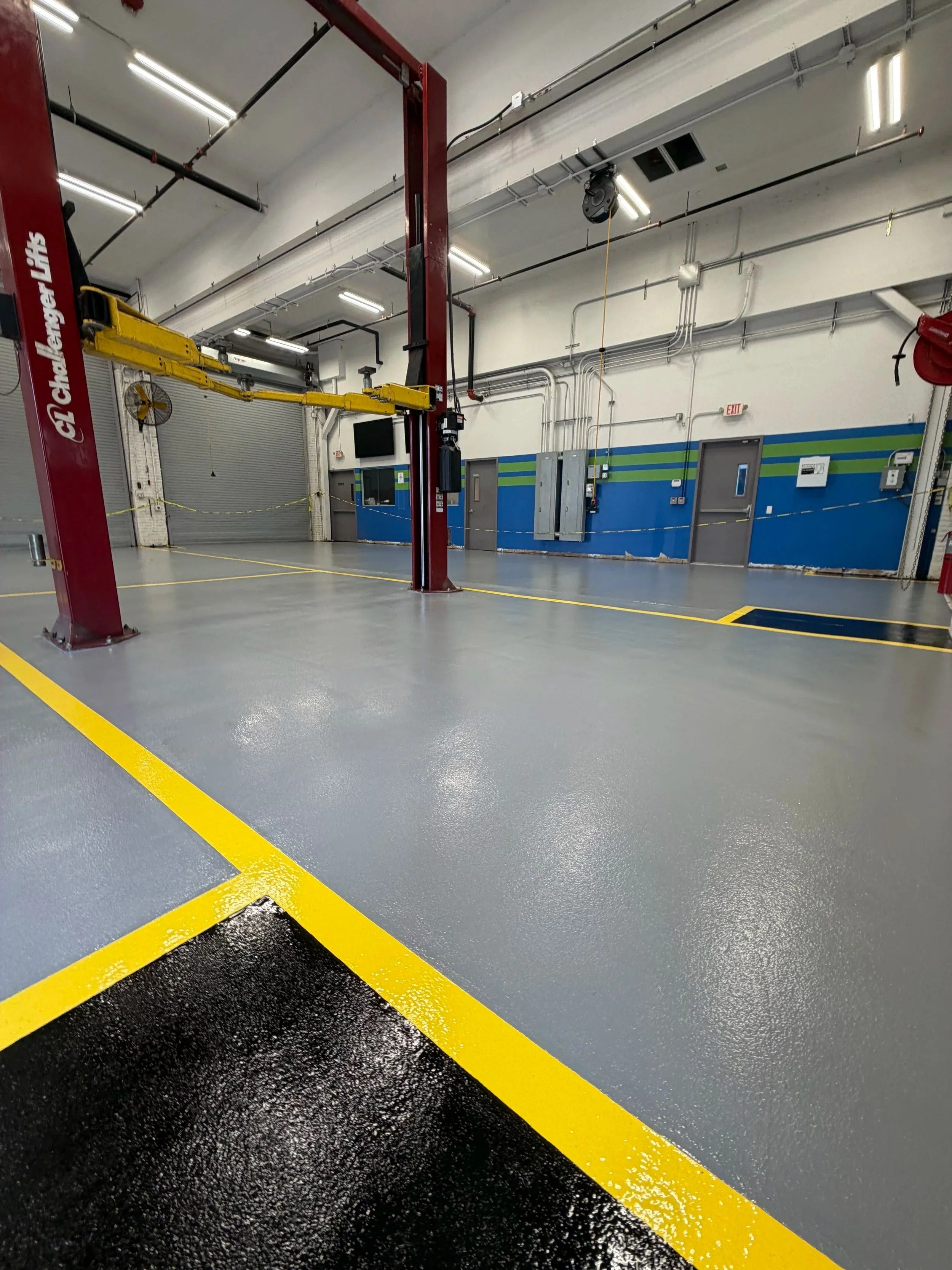 Empty industrial or workshop space with a speckled gray floor, a yellow parking curb, and a wall with orange traffic cones, buckets, ladders, and tools stored along the side.
