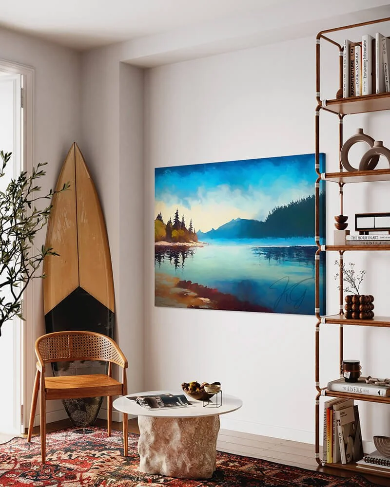 A tranquil painting of Vancouver Island, with trees, mountains, and water, hung on a wall in a cozy corner with a surfboard.