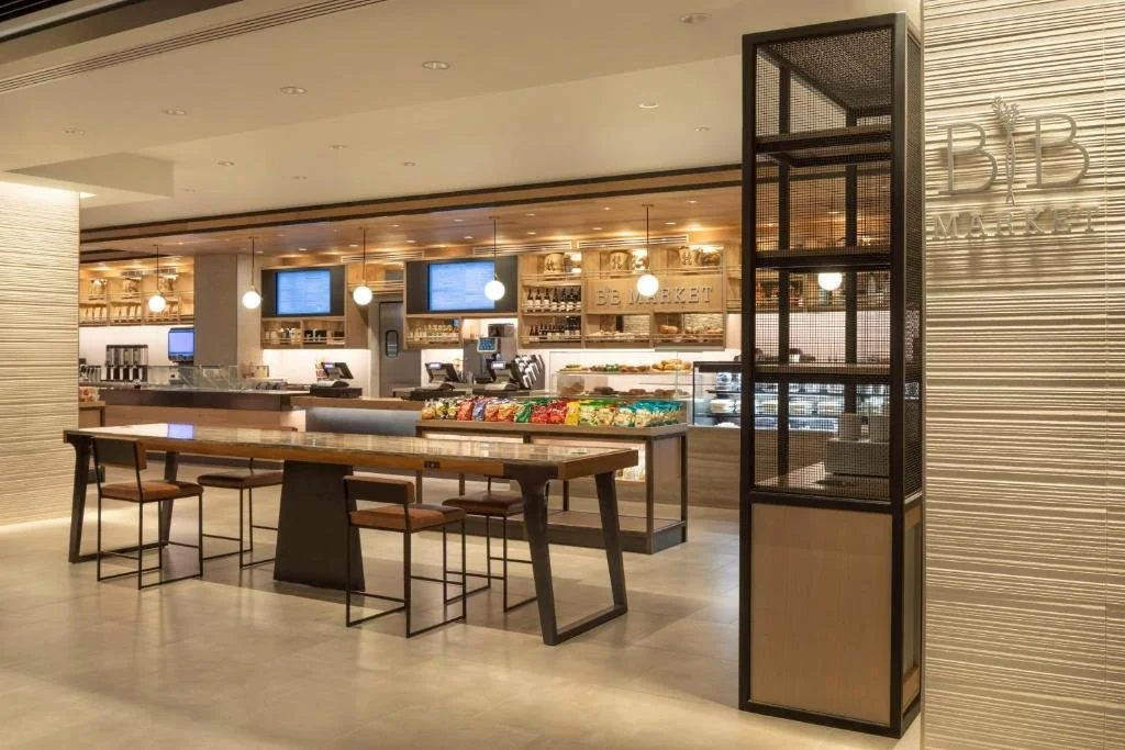Interior of a modern market or cafe with a long table, snack display, and digital menu screens.