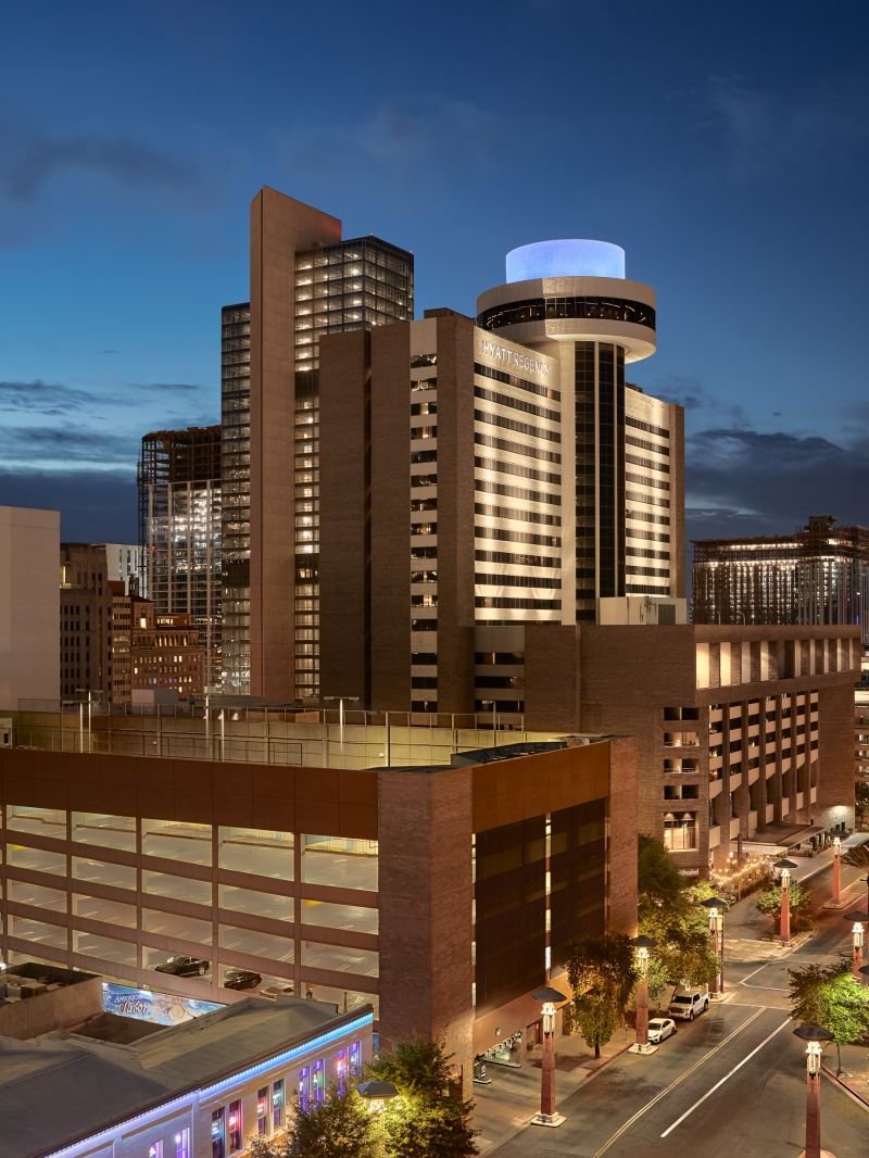 A cityscape at dusk featuring a modern high-rise hotel with lit windows and a round rooftop structure, surrounded by other tall buildings and a street with cars and trees.