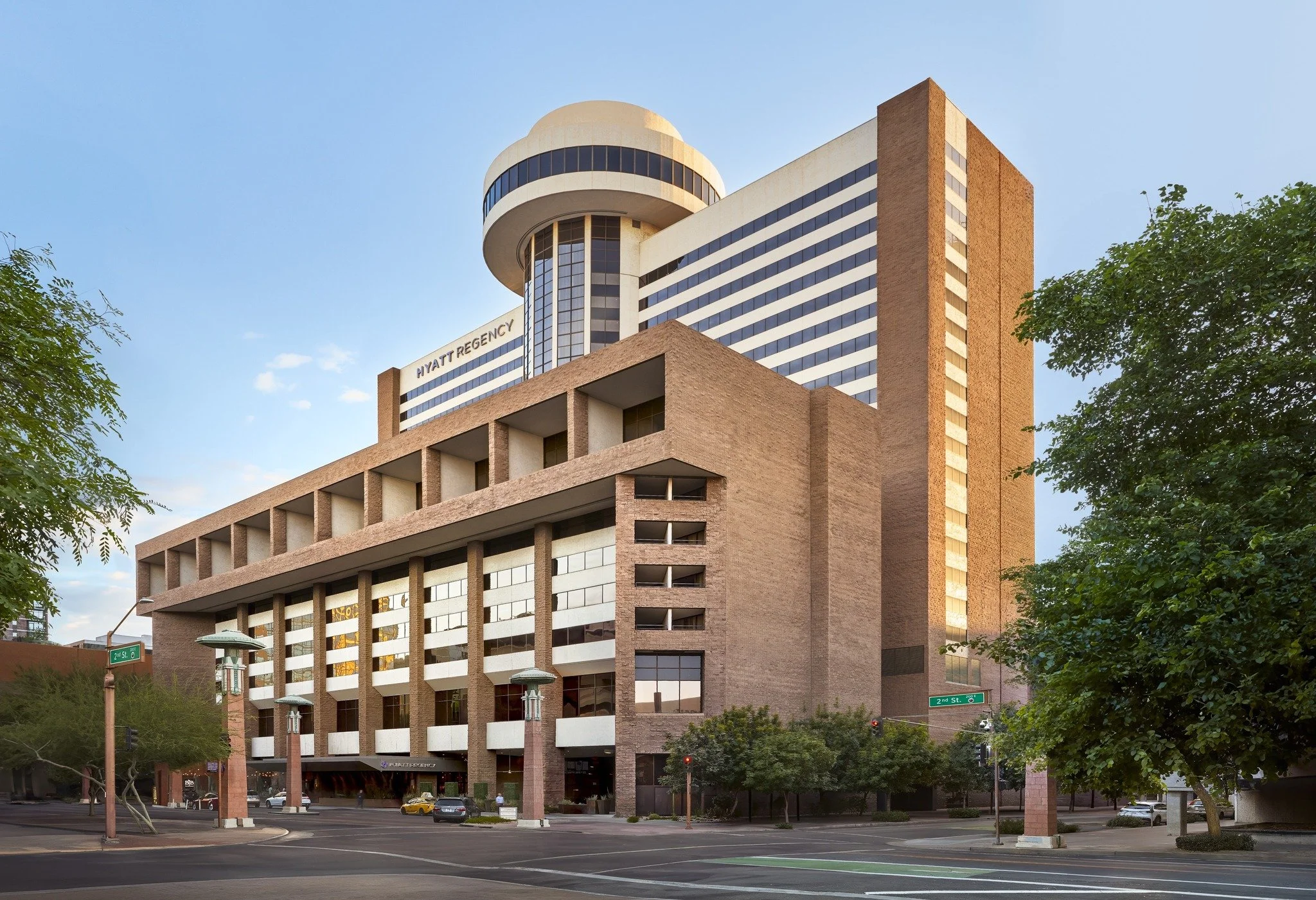 A modern multi-story hotel building with the sign "Hyatt Regency" on the upper part, featuring a distinctive rounded tower with black windows, and a brick facade with large windows. The hotel is situated on a city street corner with trees, street signs, and parked cars visible in the foreground.