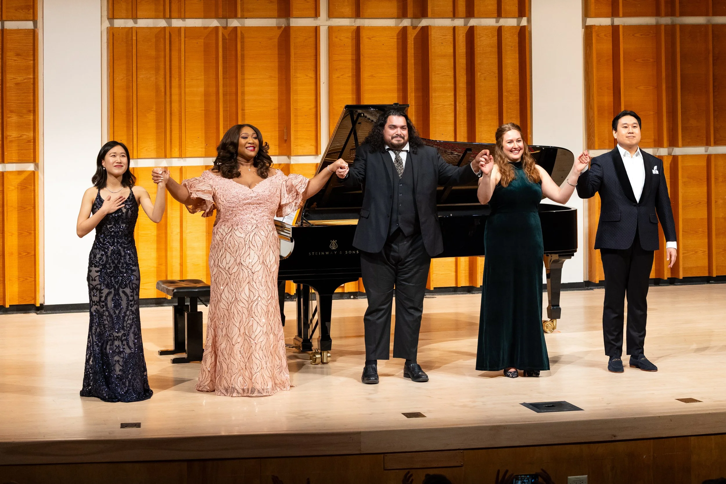 Six performers in formal attire holding hands and bowing on stage with a black Steinway & Sons grand piano behind them, set against a wooden acoustic panel background.