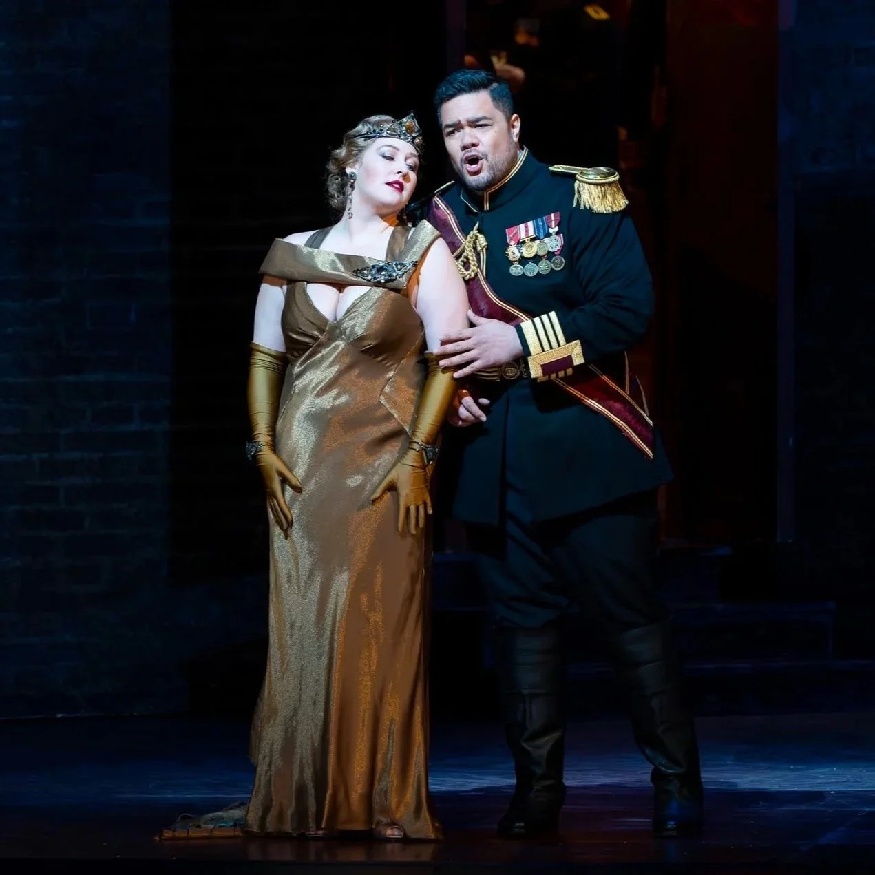 Two actors dressed in historical costumes performing on stage. The woman wears a gold gown, and the man is in a military uniform with medals.