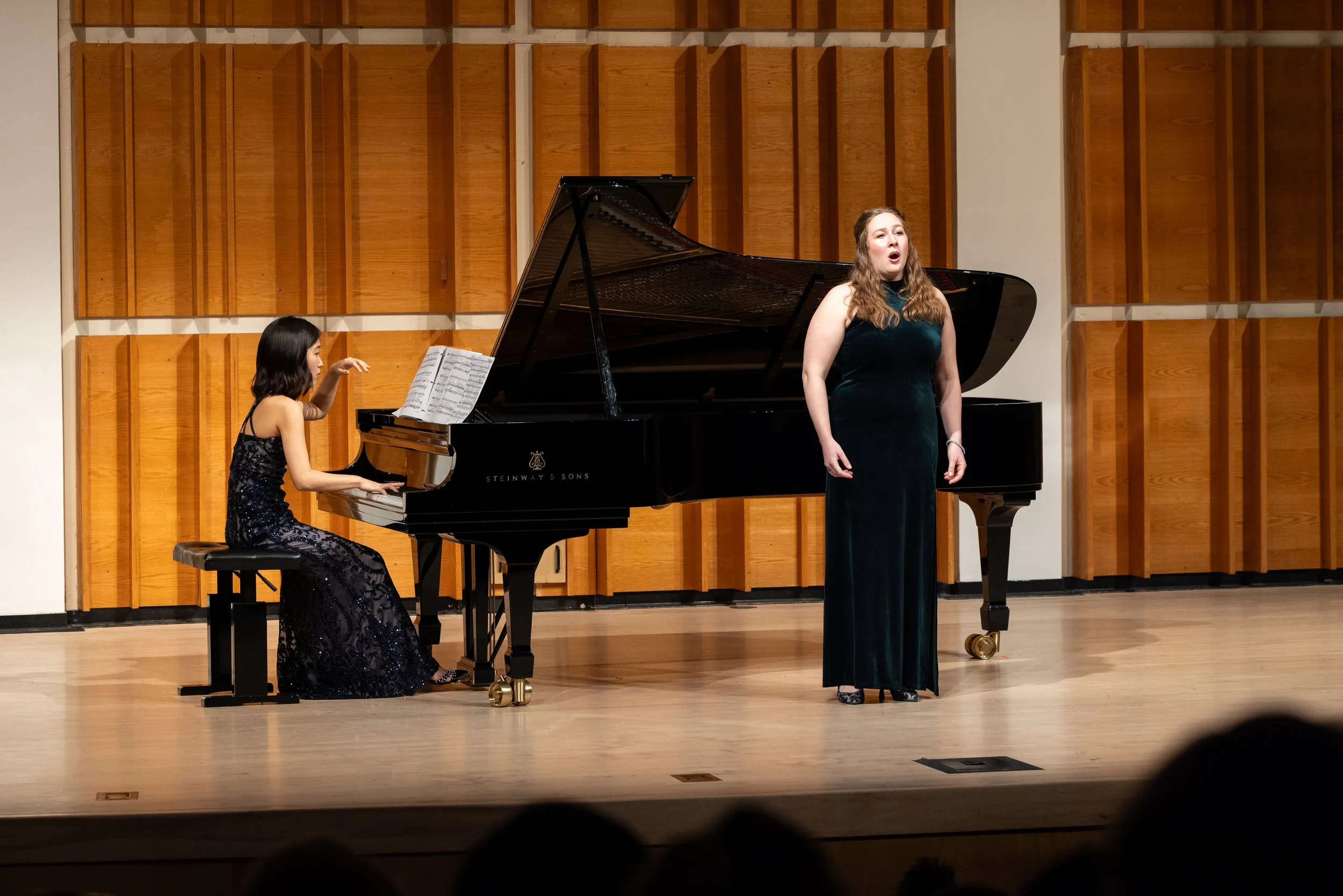 A woman singing on stage beside a woman playing the piano during a performance, with a wooden panel background.