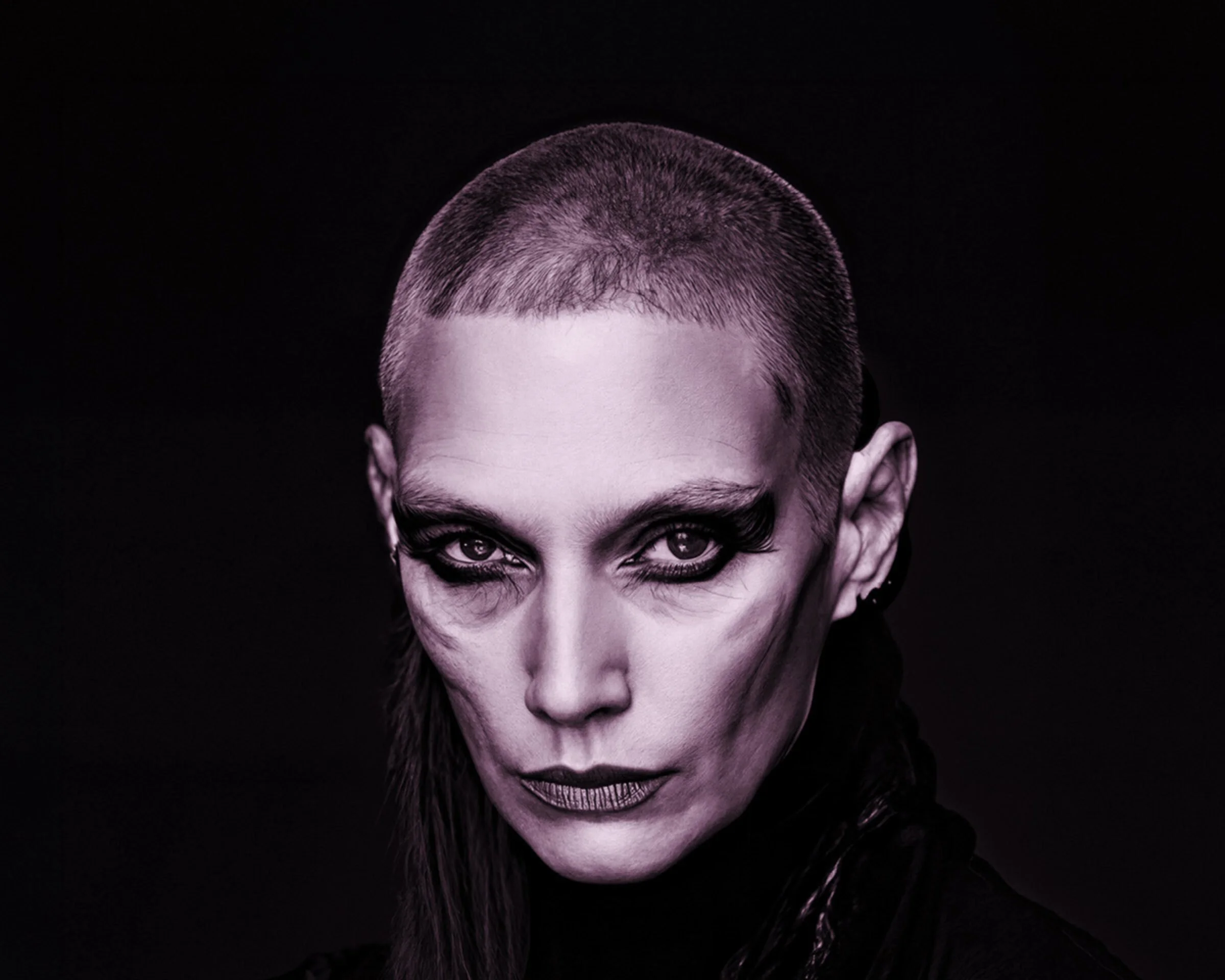 Close-up of a woman with bold makeup and a shaved hairstyle, looking directly at the camera against a black background.