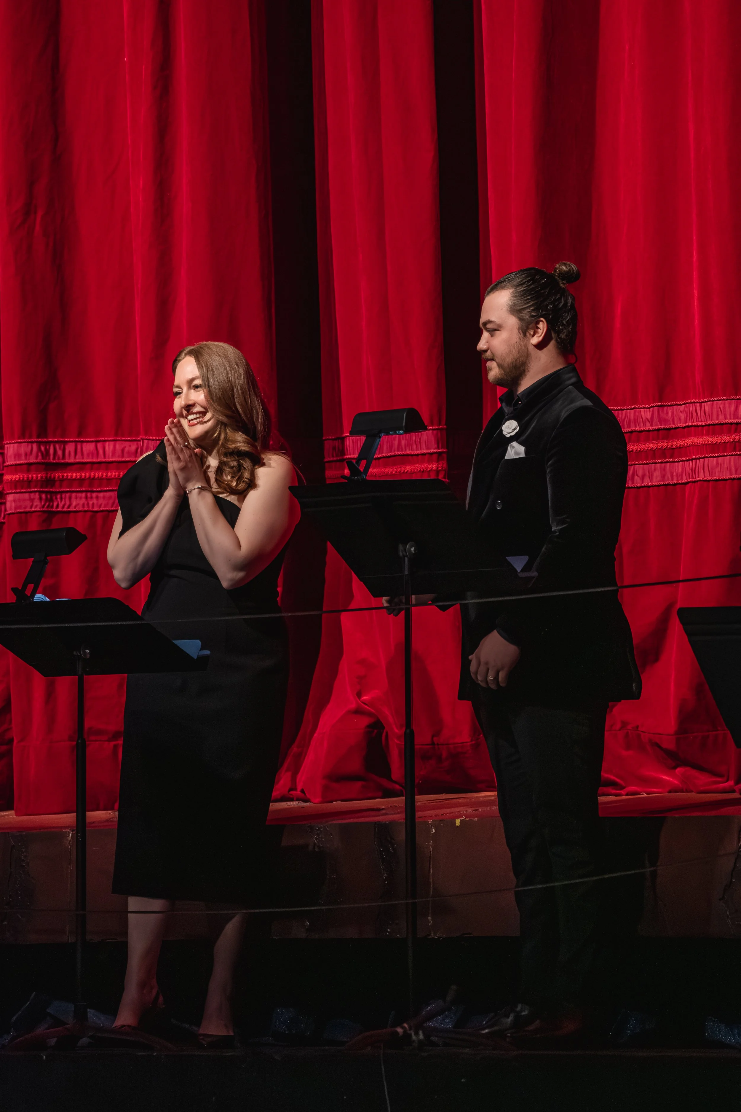 A woman with long hair wearing a black dress and a man with a beard and tied-back hair wearing a black suit on stage with red curtains in the background. The woman is smiling with her hands clasped near her face, and the man appears to be speaking or singing at a music stand.