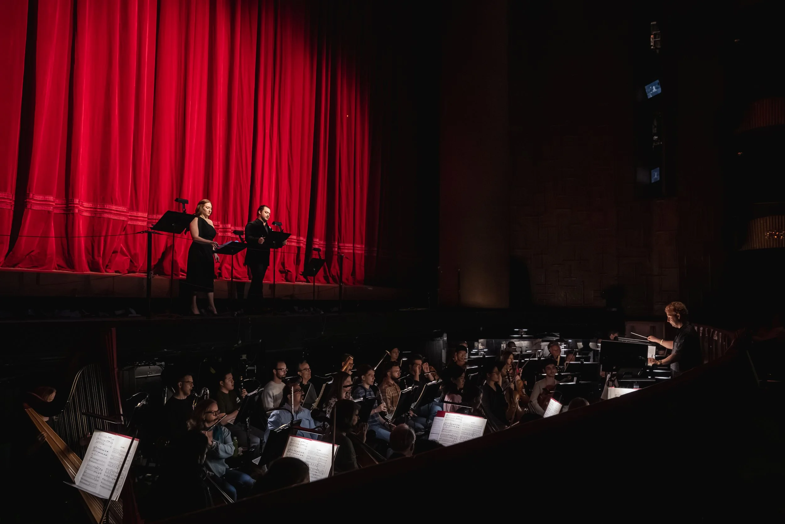 Orchestra performing on stage with red velvet curtains, with two conductors and musicians with sheet music focused on playing their instruments.