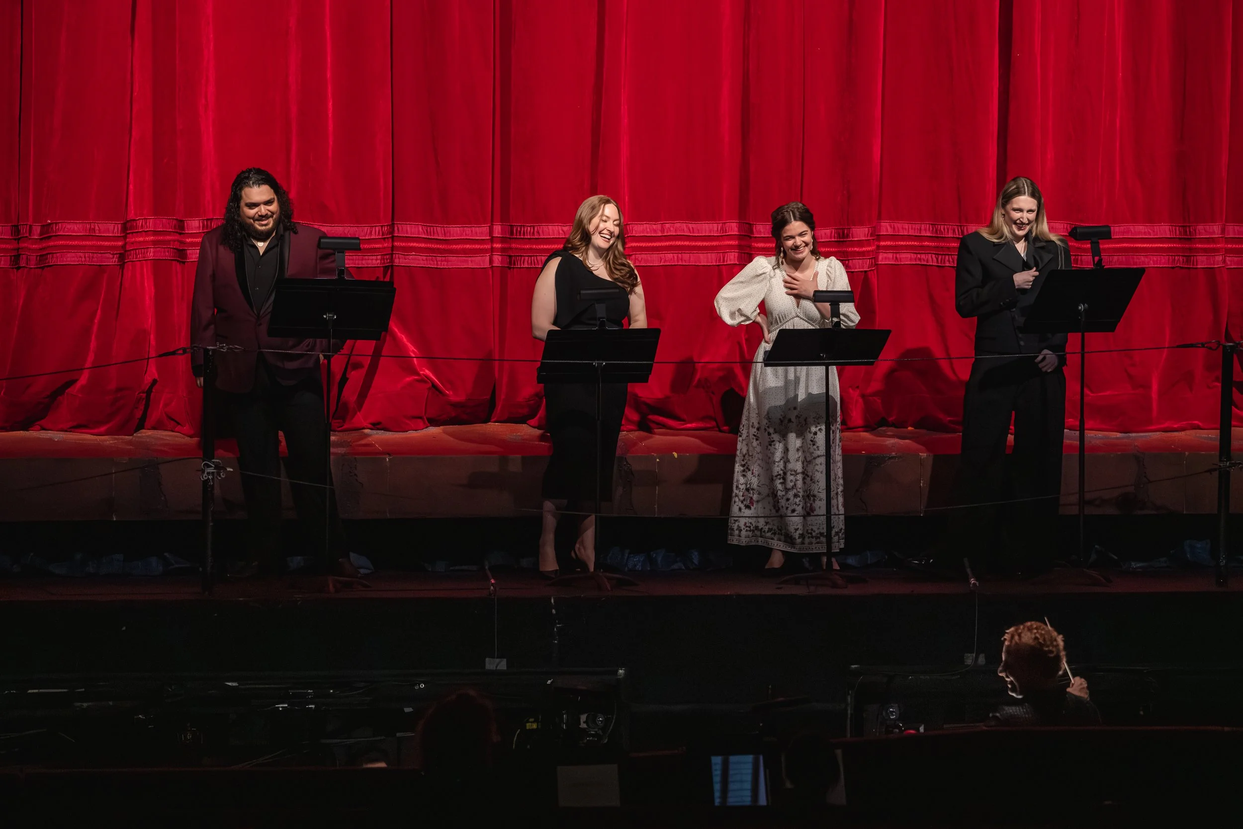 Four performers standing on stage in front of a red curtain, smiling and engaging with the audience. One person is reading from a script, while the others are smiling or with one hand on their chest.