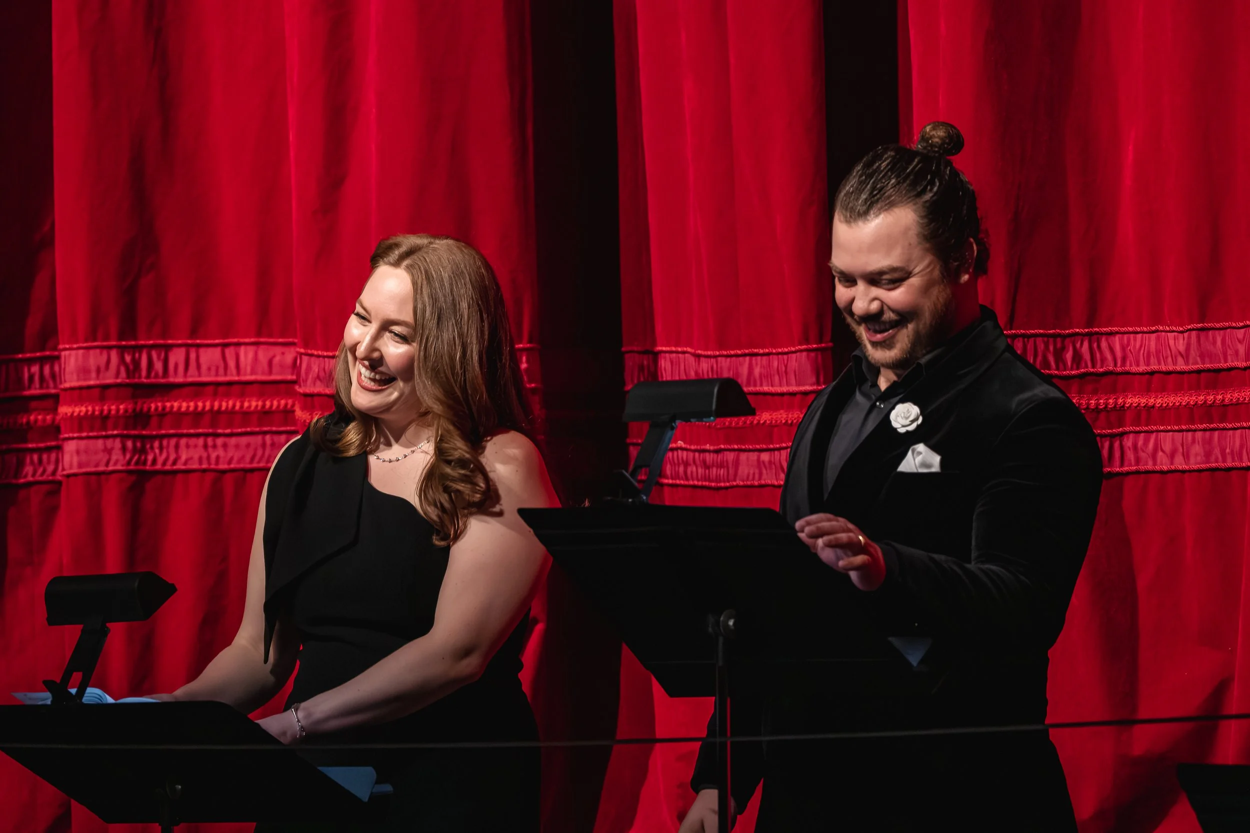 Two smiling musicians, a woman and a man, performing on stage with late red curtains in the background. The woman is at a keyboard, and the man is playing a piano, both dressed in formal attire.