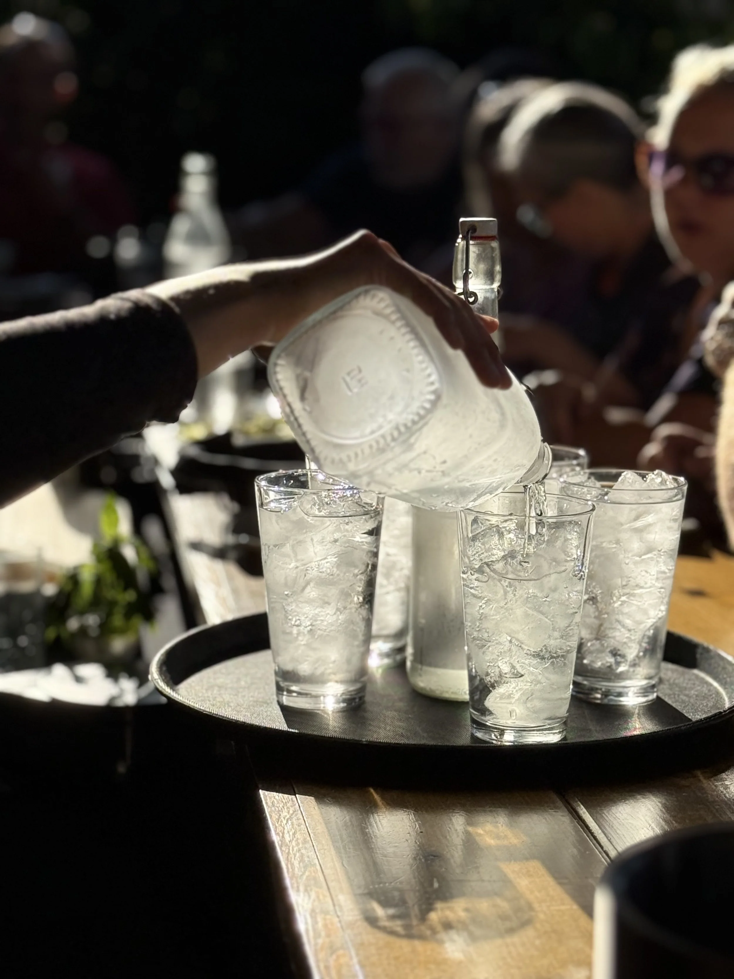 BArtender pouring cocktails, water or soda, into a glass filled with ice on a beautiful patio. In the background, several people are seated, with some wearing sunglasses, in an outdoor setting.