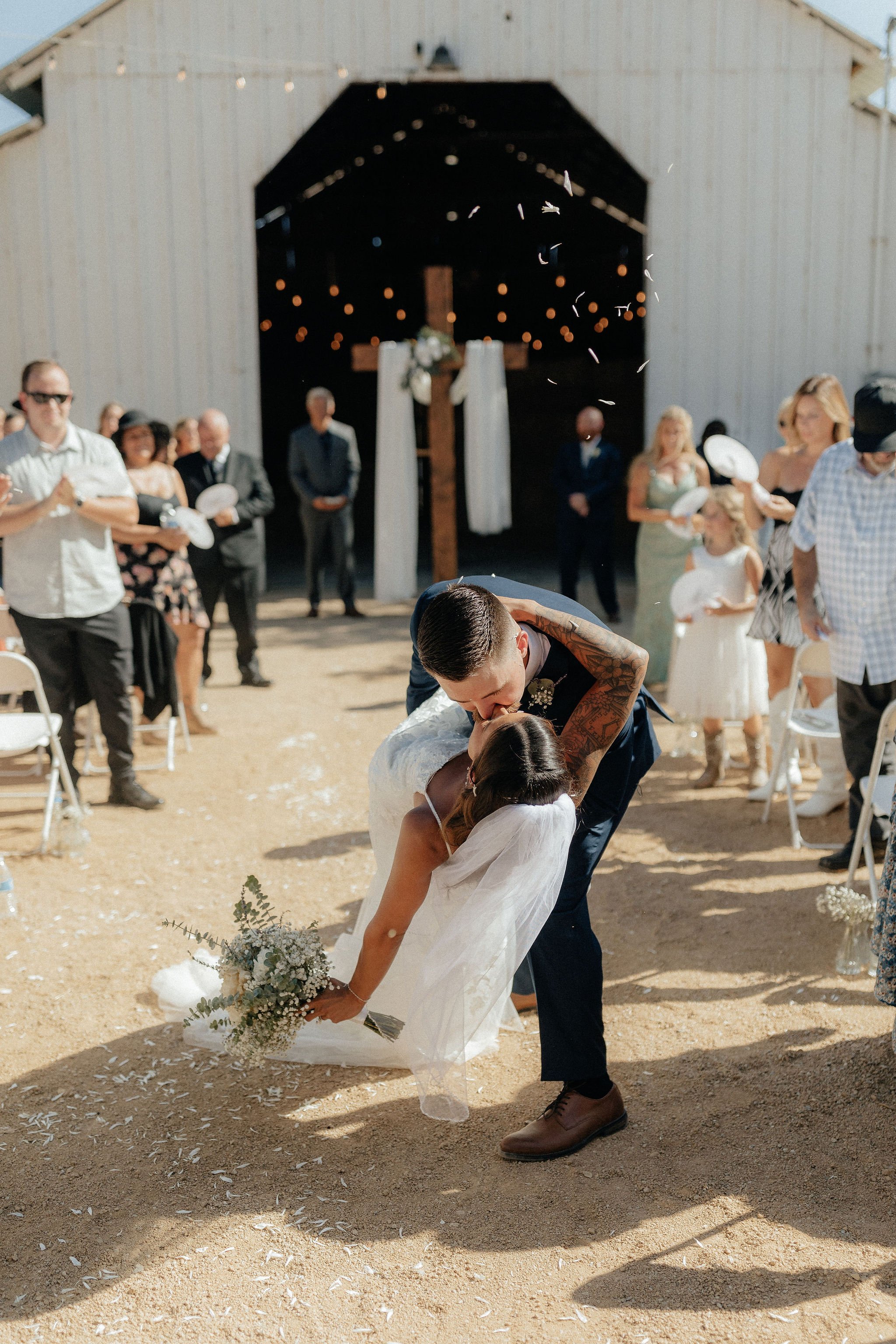 A bride and groom are kissing during their wedding ceremony outside a barn, with guests watching and a cross in the background.