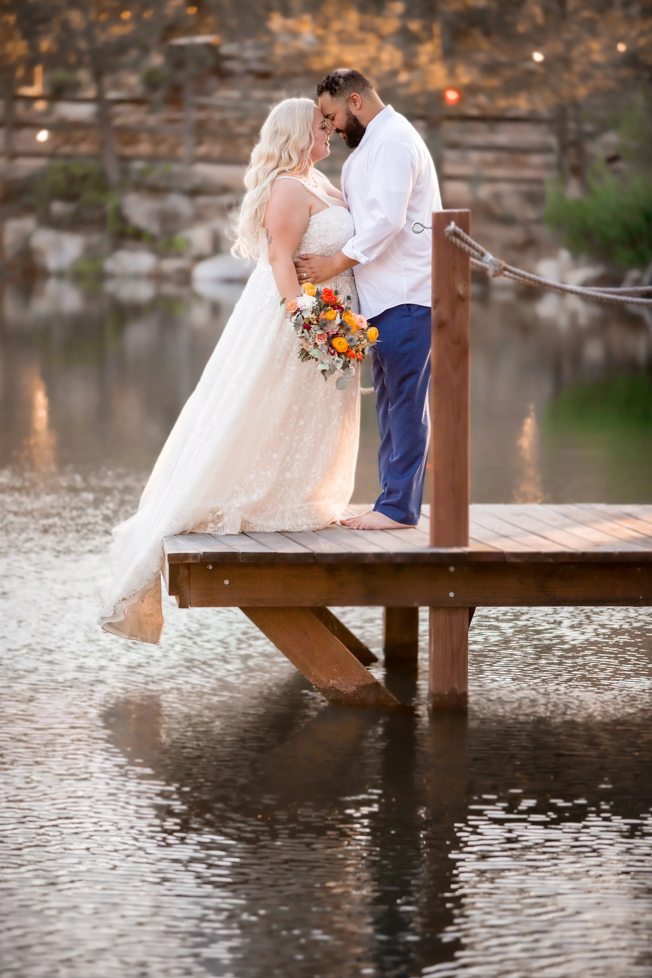 A bride and groom standing close together on a wooden dock over water, with their foreheads touching and eyes closed, during sunset. The bride is holding a colorful bouquet, and the background features blurred trees and rocky landscape.