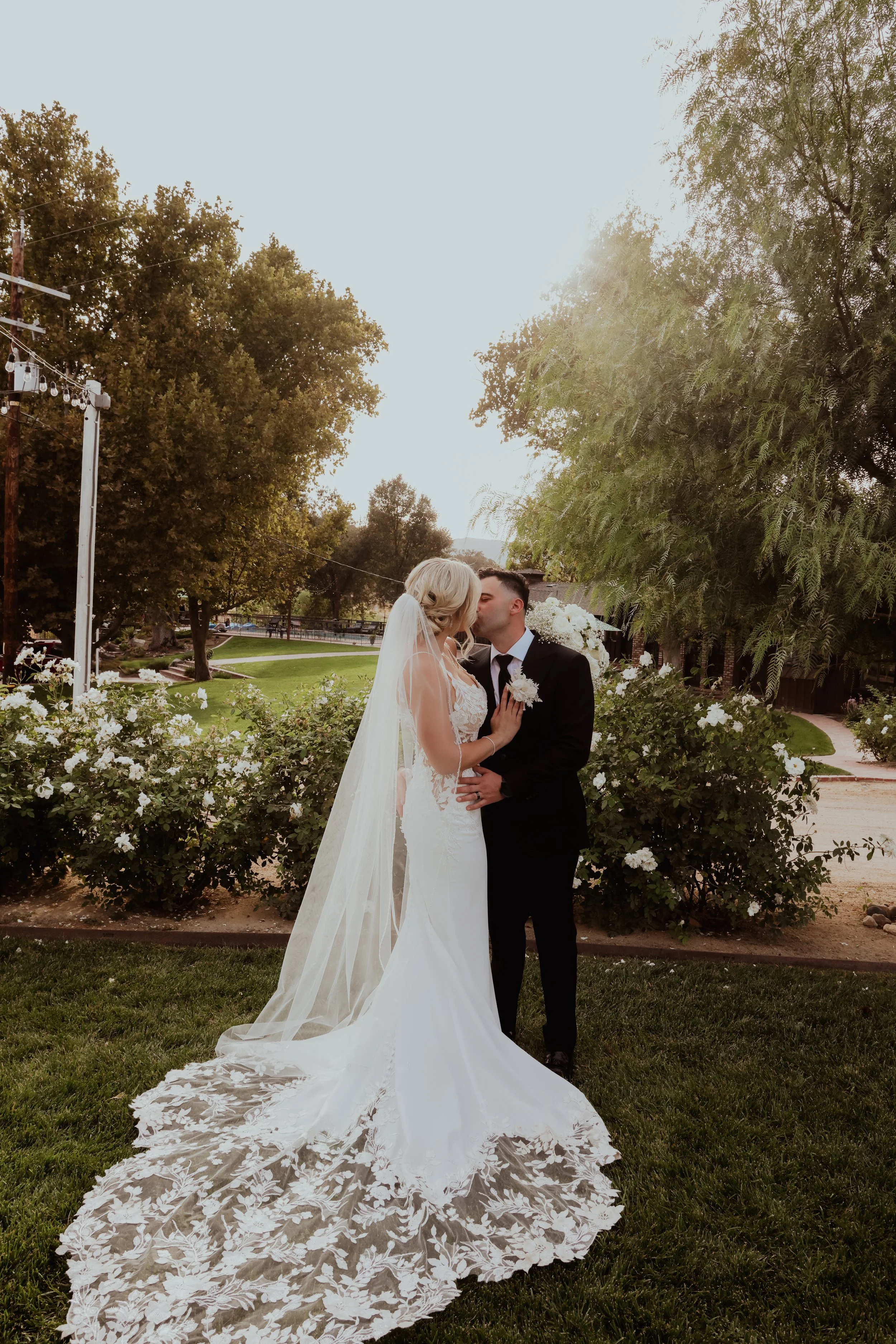 A bride and groom share a kiss in a garden during their wedding, with lush green trees and a blue sky in the background.