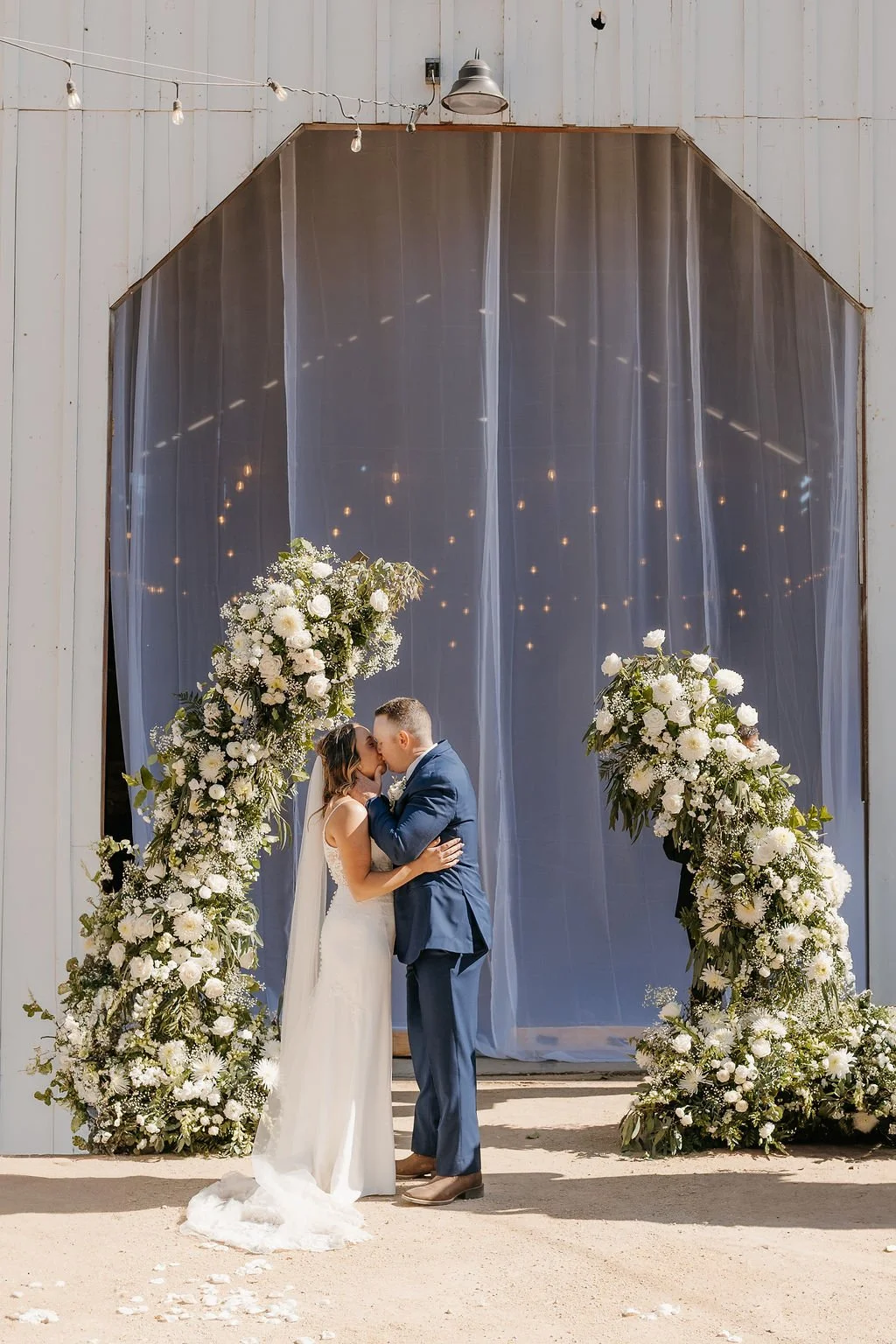 A bride and groom share a kiss during their wedding ceremony in front of a floral arch with white flowers and greenery, set against a rustic backdrop with fairy lights.