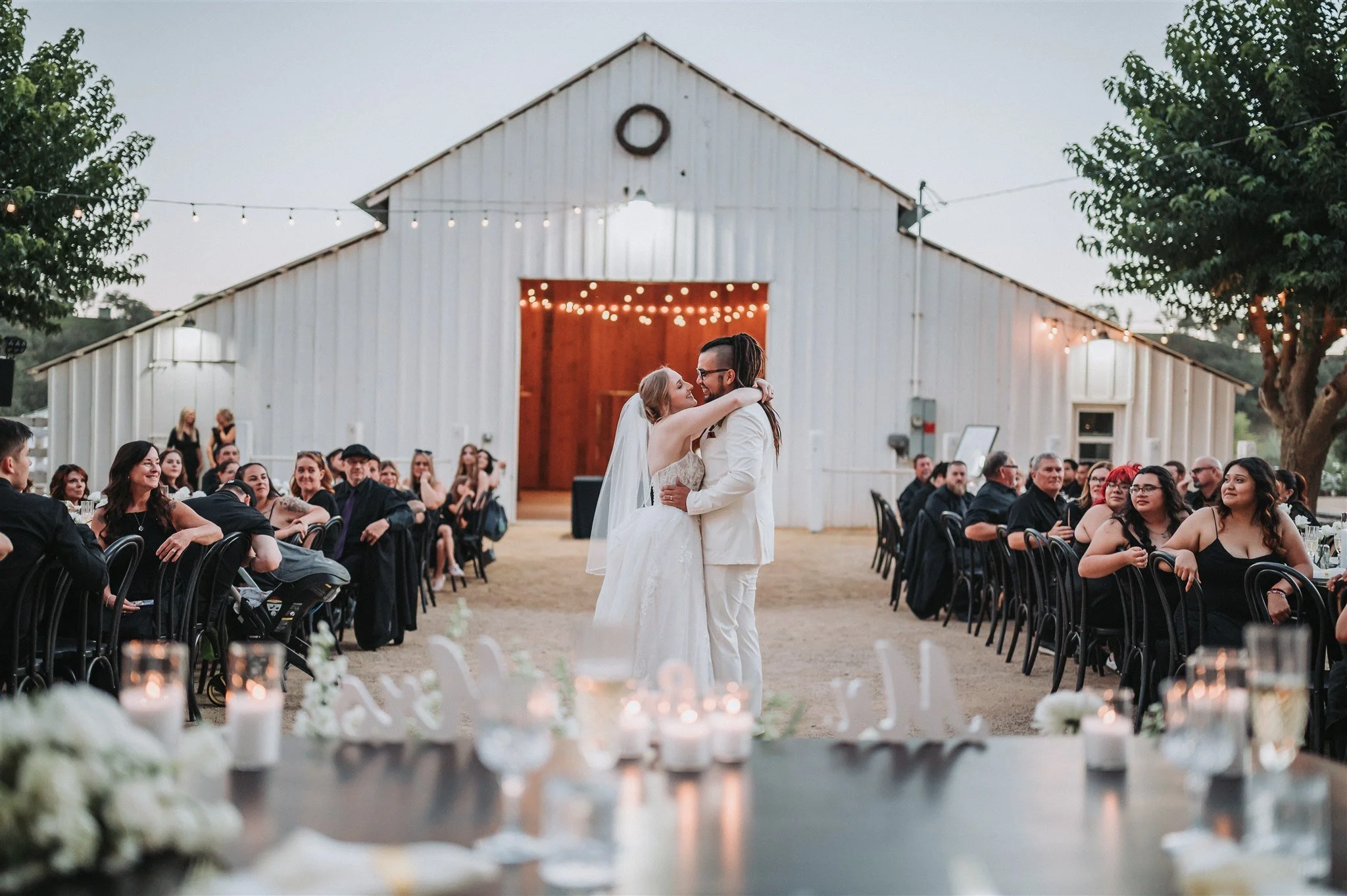 A newlywed couple dancing at an outdoor wedding reception in front of a rustic barn, with guests seated at tables on either side, string lights overhead, and candles in the foreground.