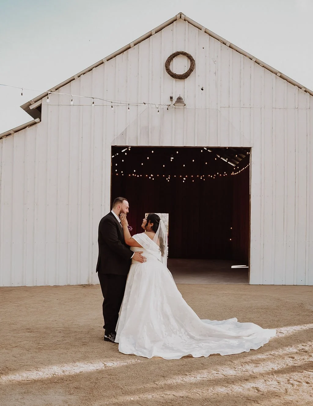 A bride and groom in wedding attire standing inside a rustic barn, affectionately touching each other's faces, with string lights hanging inside and a wreath on the barn's exterior.