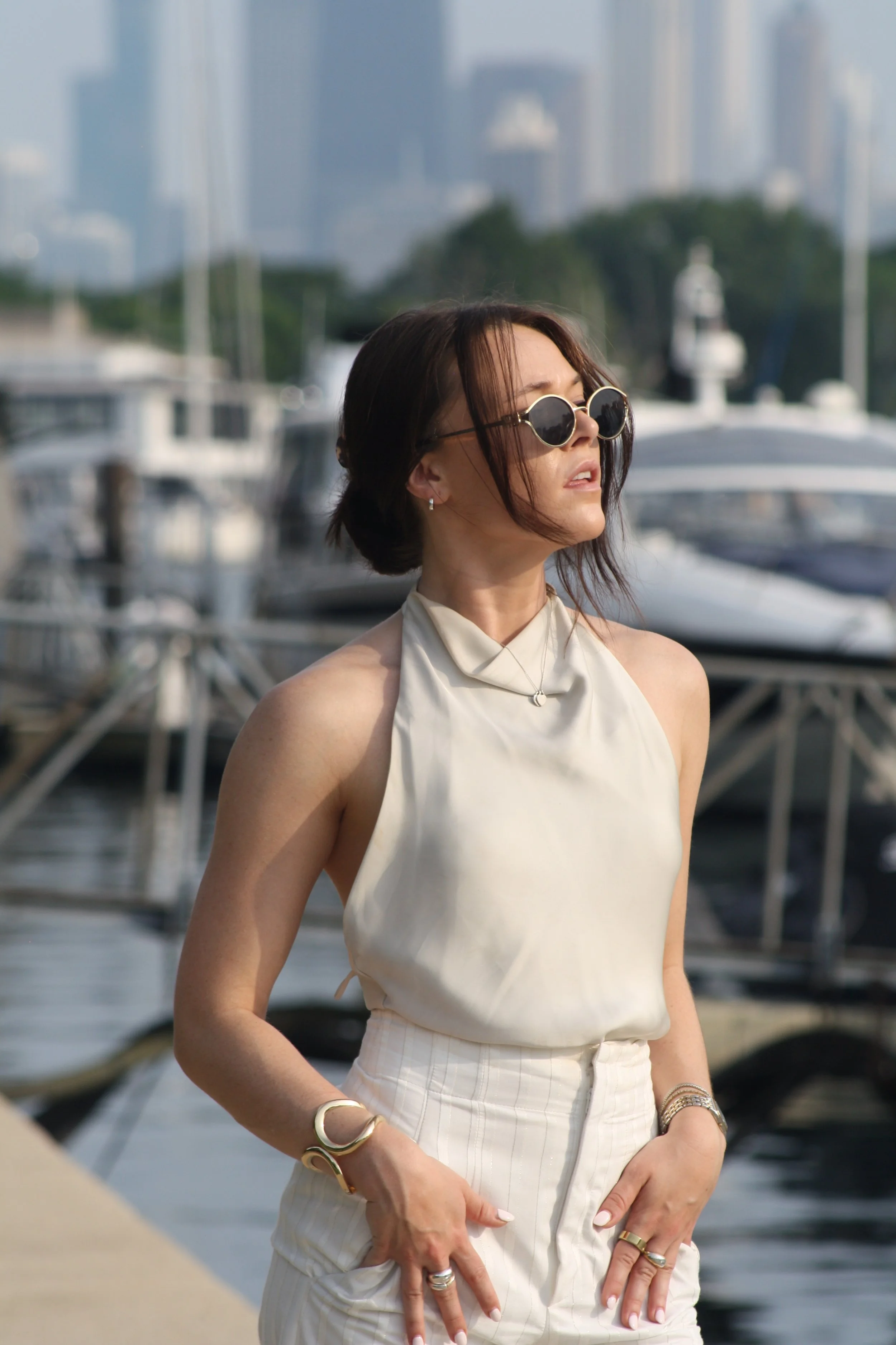 Woman wearing sunglasses, a sleeveless cream-colored silk top, and cream high-waisted trousers, standing near a marina with boats and yachts in the background.