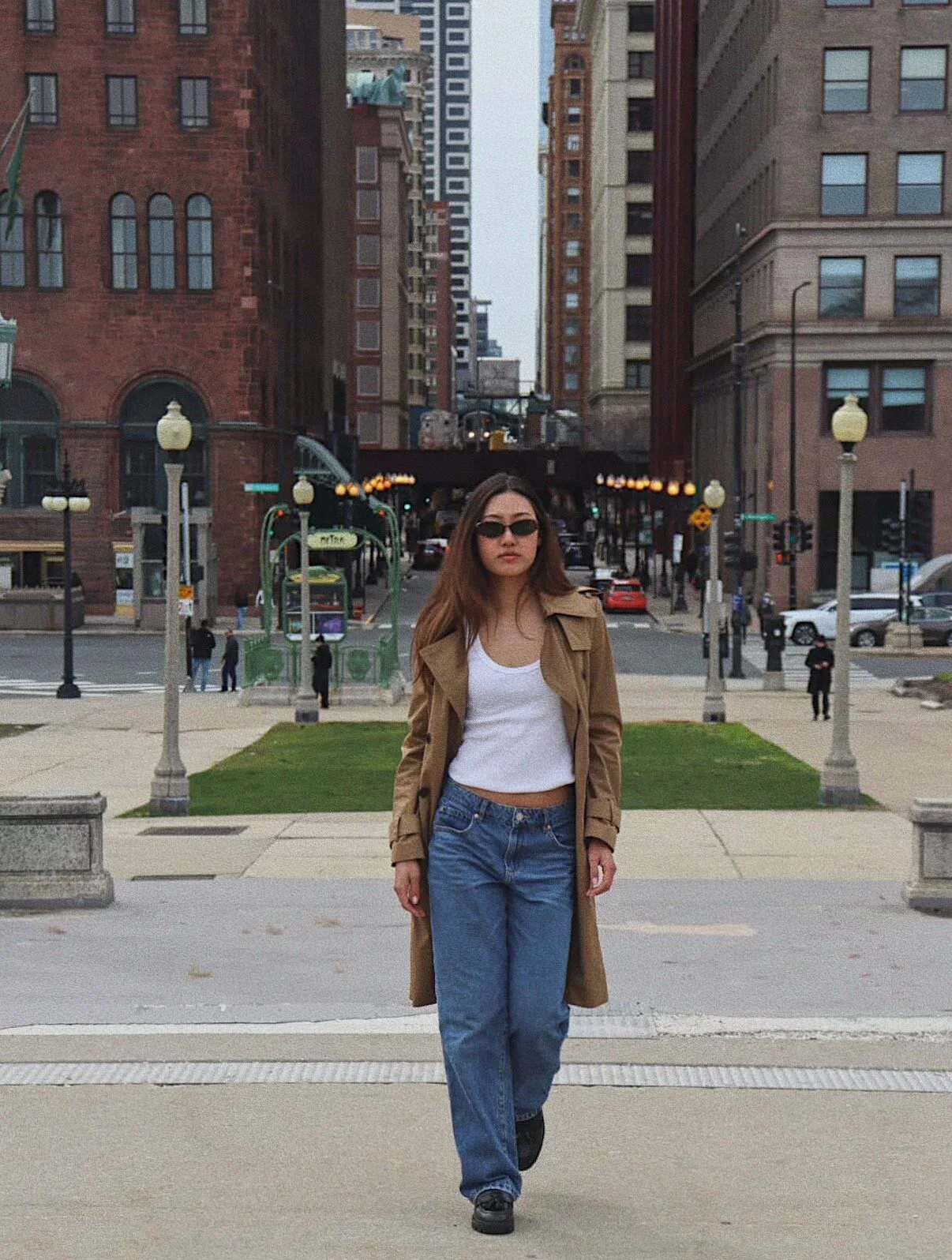 A woman walking on the sidewalk in an urban city, wearing sunglasses, a tan trench coat, a white top, and blue jeans, with tall buildings and a street with cars in the background.