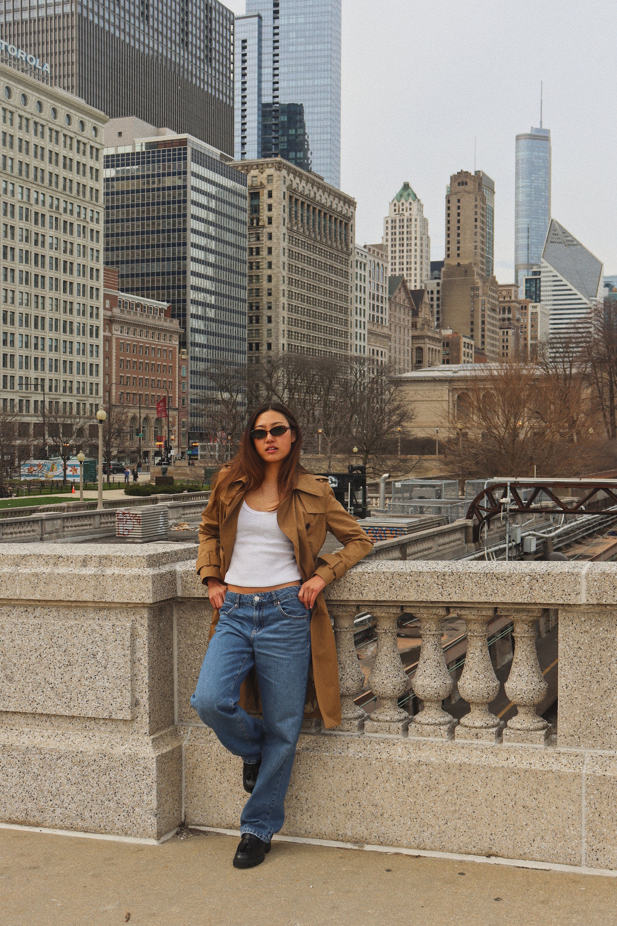 A young woman with long brown hair wearing sunglasses, a white crop top, blue jeans, black shoes, and a tan trench coat, leaning against a stone balcony railing in an urban cityscape with tall modern and historic buildings.