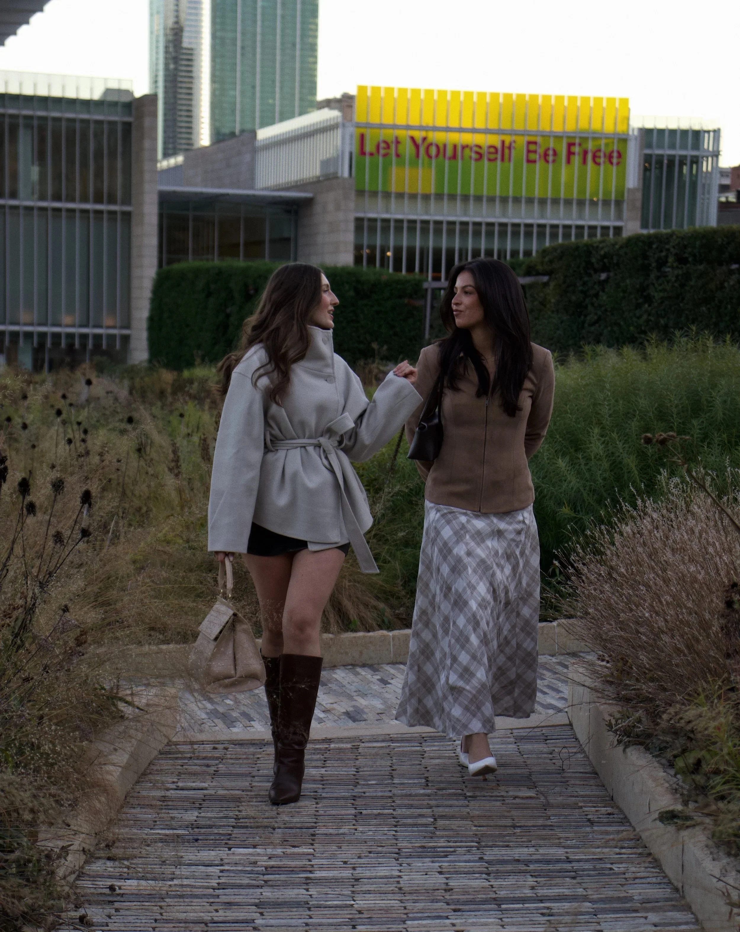 Two women walking on a wooden pathway in an urban park, with modern buildings and a sign that reads "Let Yourself Be Free" in the background.