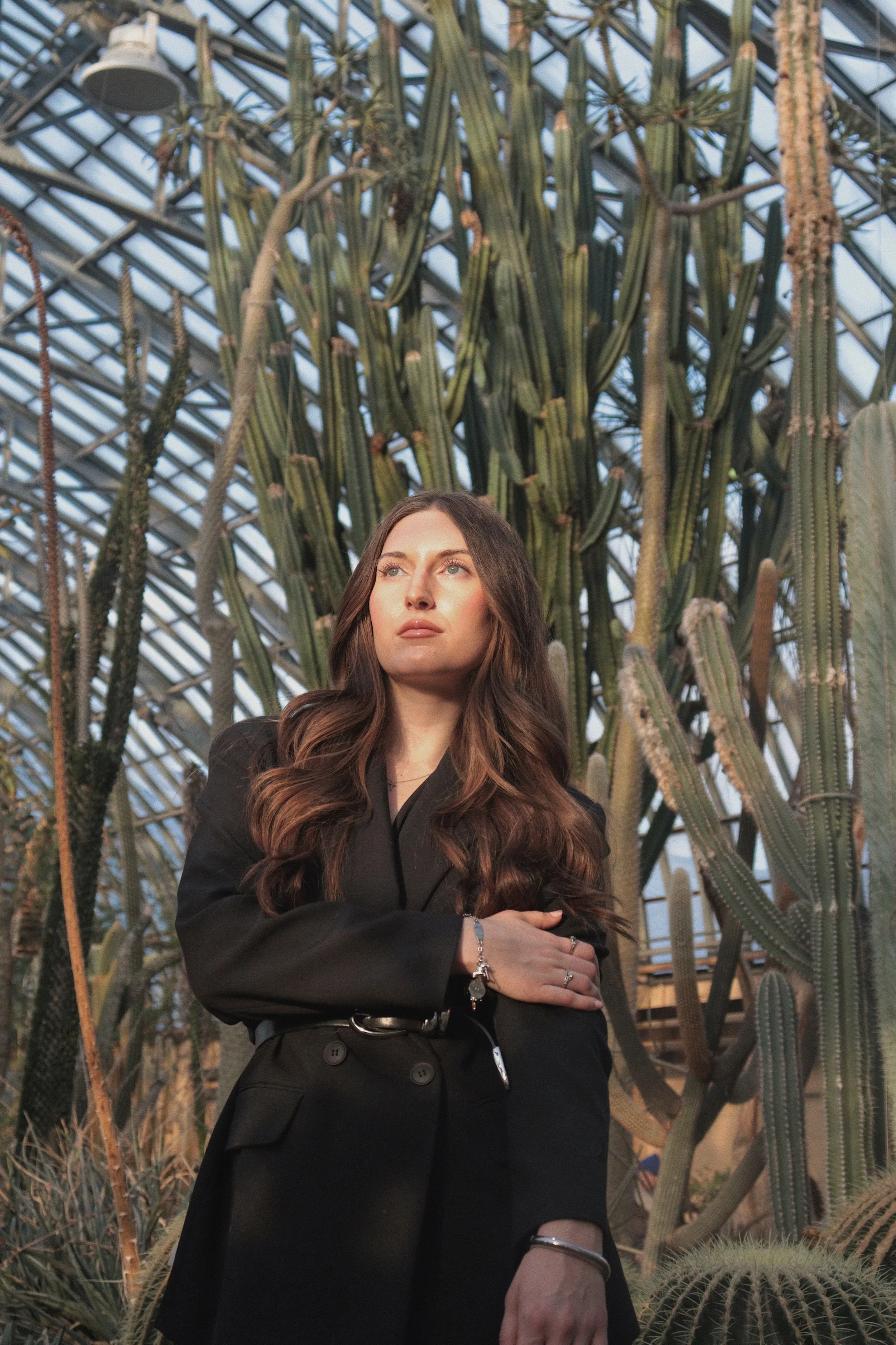 A woman with long, wavy brown hair and a serious expression stands in front of tall cacti inside a greenhouse or conservatory with a glass ceiling. She is dressed in a black blazer and accessorized with rings, a bracelet, and a watch.