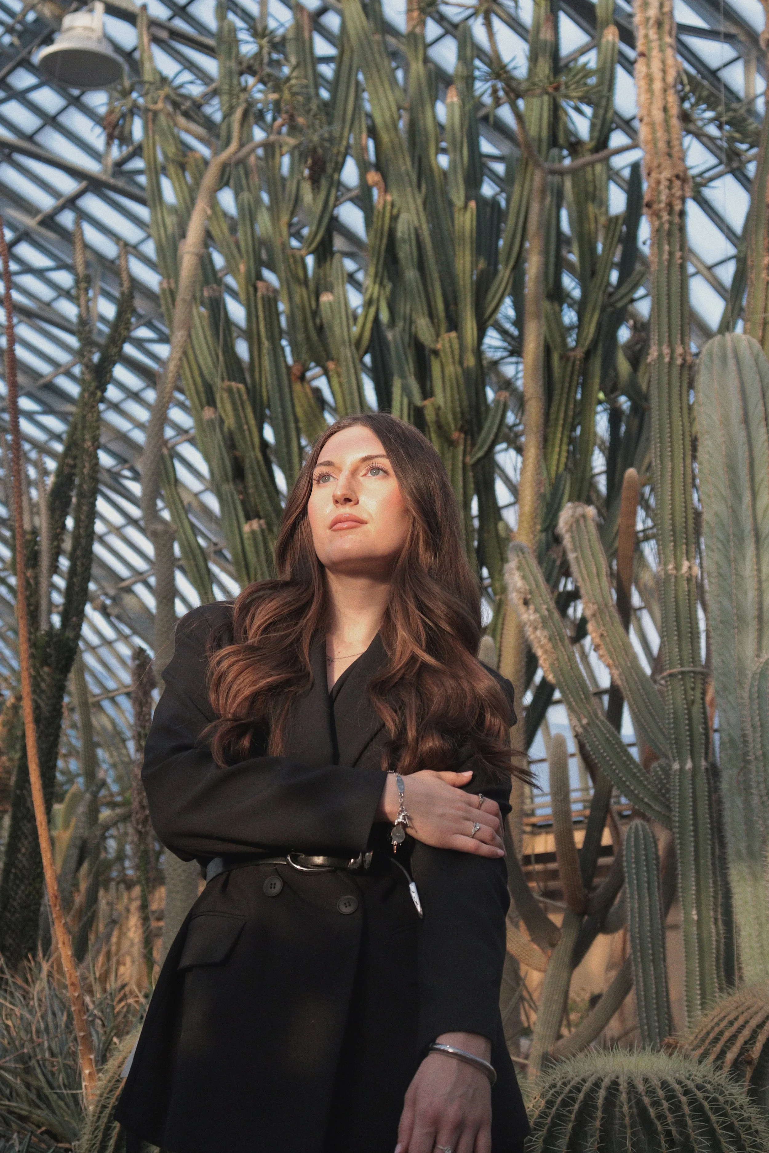 A woman with long brown hair wearing a black blazer and multiple rings, standing in front of tall cacti inside a greenhouse or botanical garden.