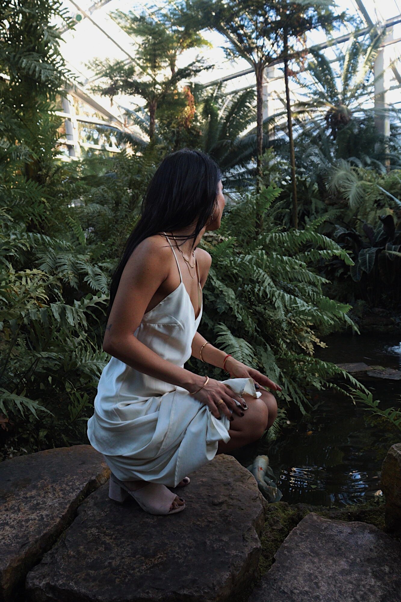 Woman in a white dress crouching on a rock by a pond surrounded by lush green plants in a greenhouse or tropical setting.
