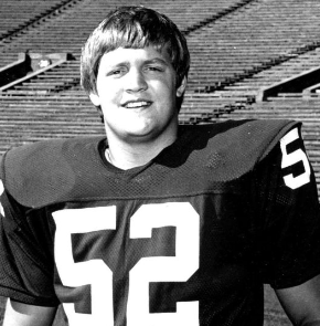 Black and white photo of a young football player wearing a jersey with the number 52, standing on a football field with empty bleachers in the background.