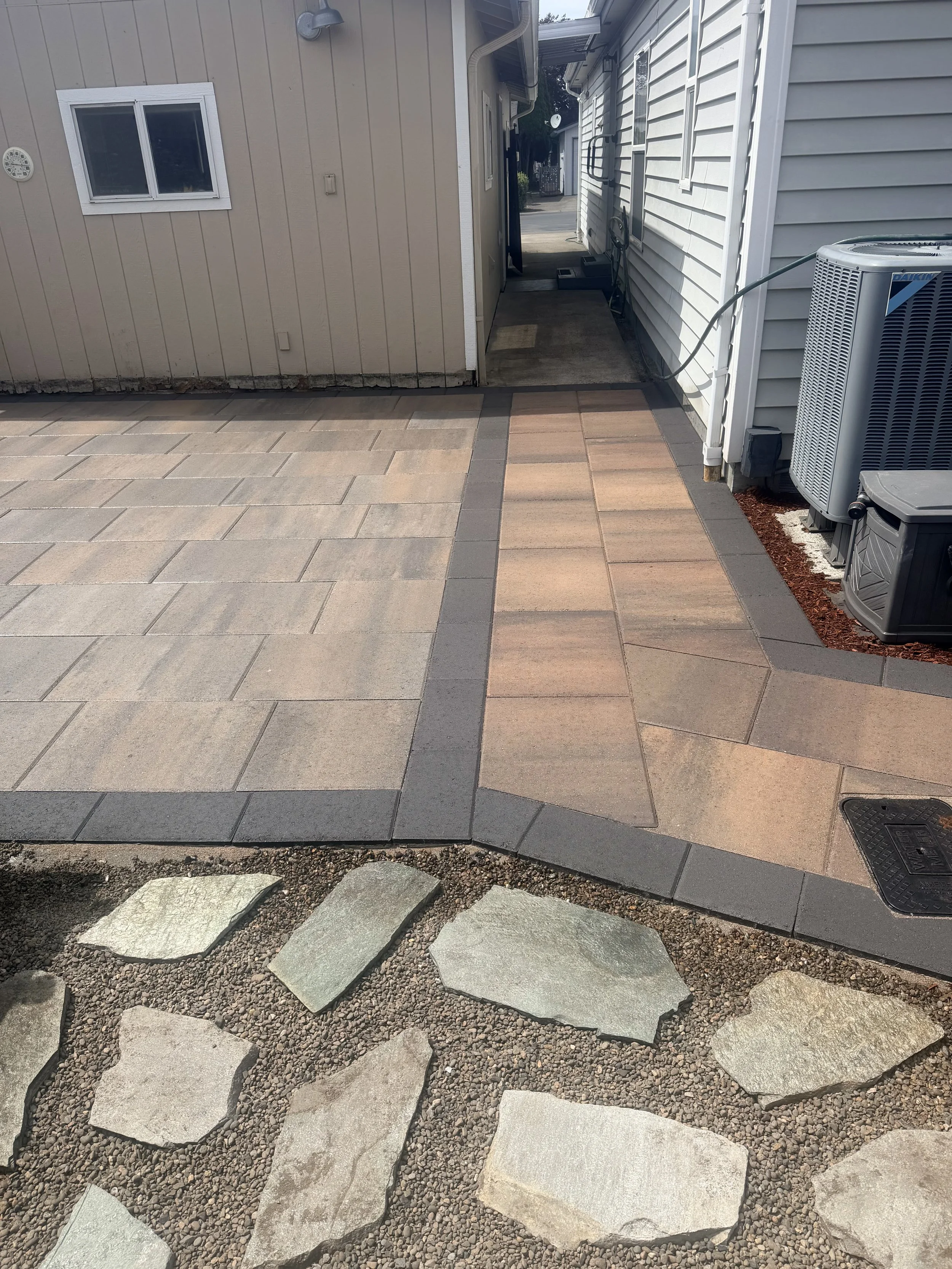 View of a backyard with a paved walkway and a stone pathway under construction. The paved area has rectangular tiles in shades of beige, gray, and reddish-brown, separated by dark gray grout lines. The stone pathway at the bottom features large, flat