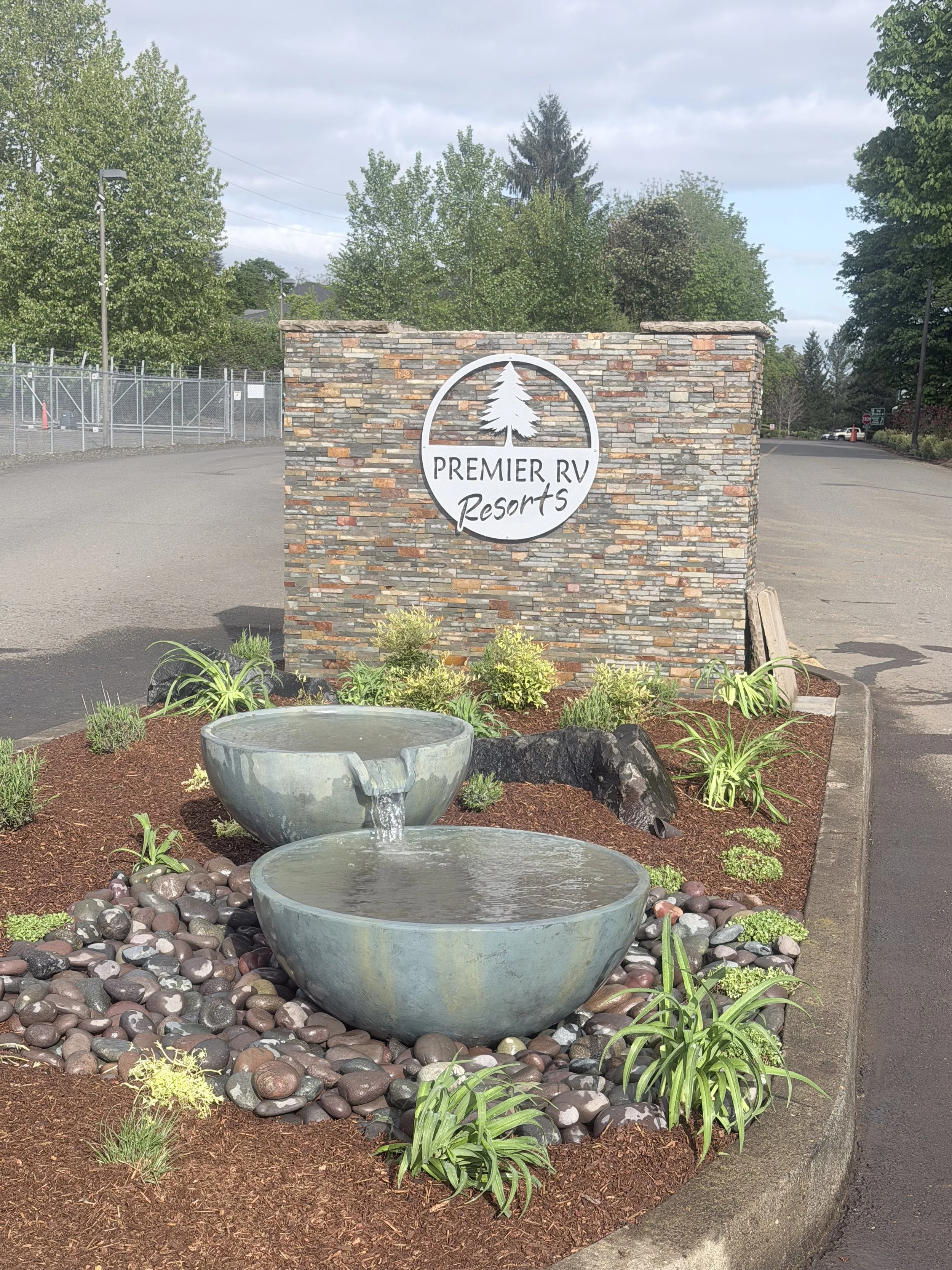 Stone sign with a white circular logo featuring a pine tree and the text 'Premier RV Resorts' in black, mounted on a brick wall. In front of the sign are two large decorative water fountains surrounded by small plants, rocks, and mulch. There are tre