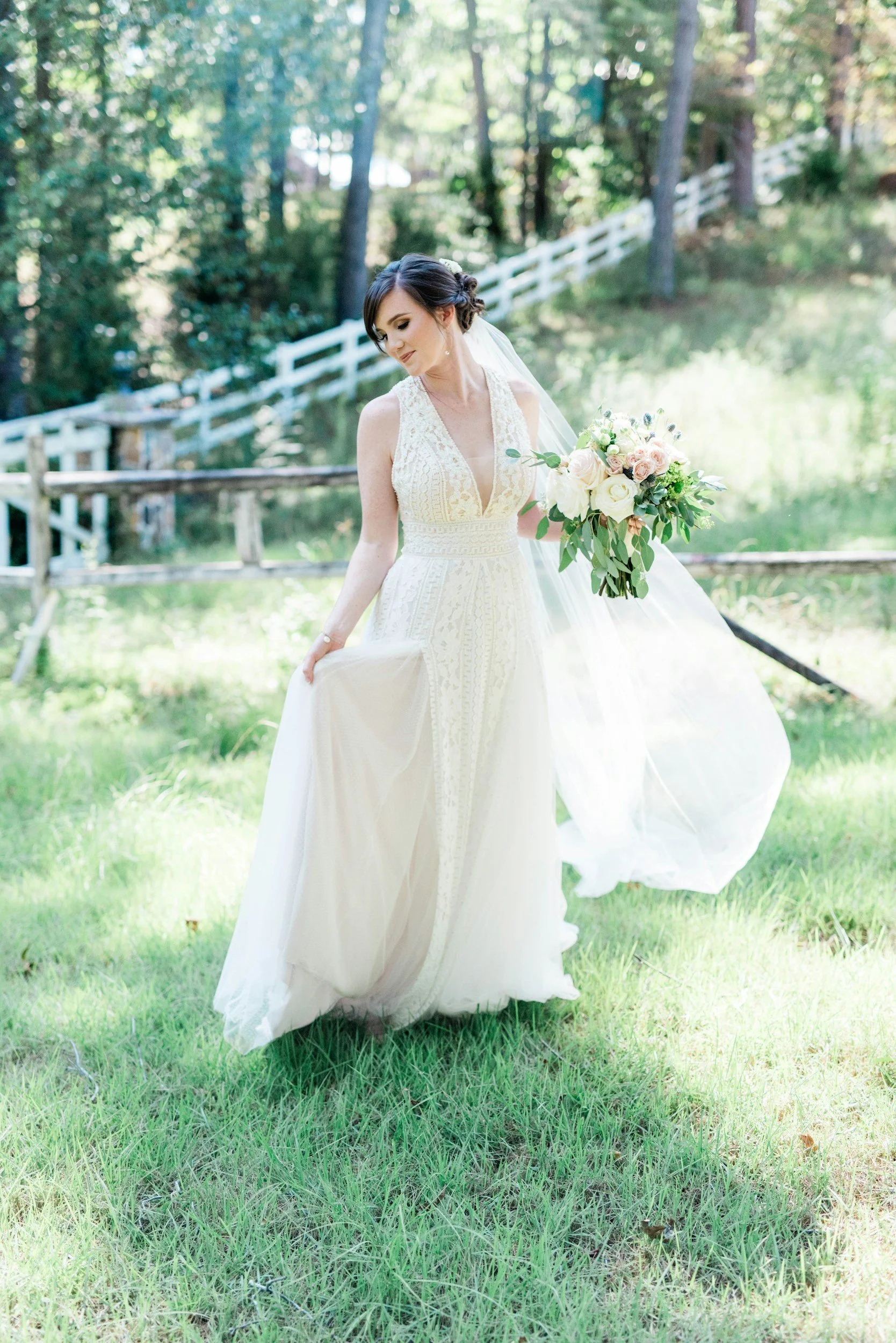 A bride in a white lace wedding dress holding a bouquet of white and pink flowers, standing outdoors on green grass with trees and a wooden fence in the background.