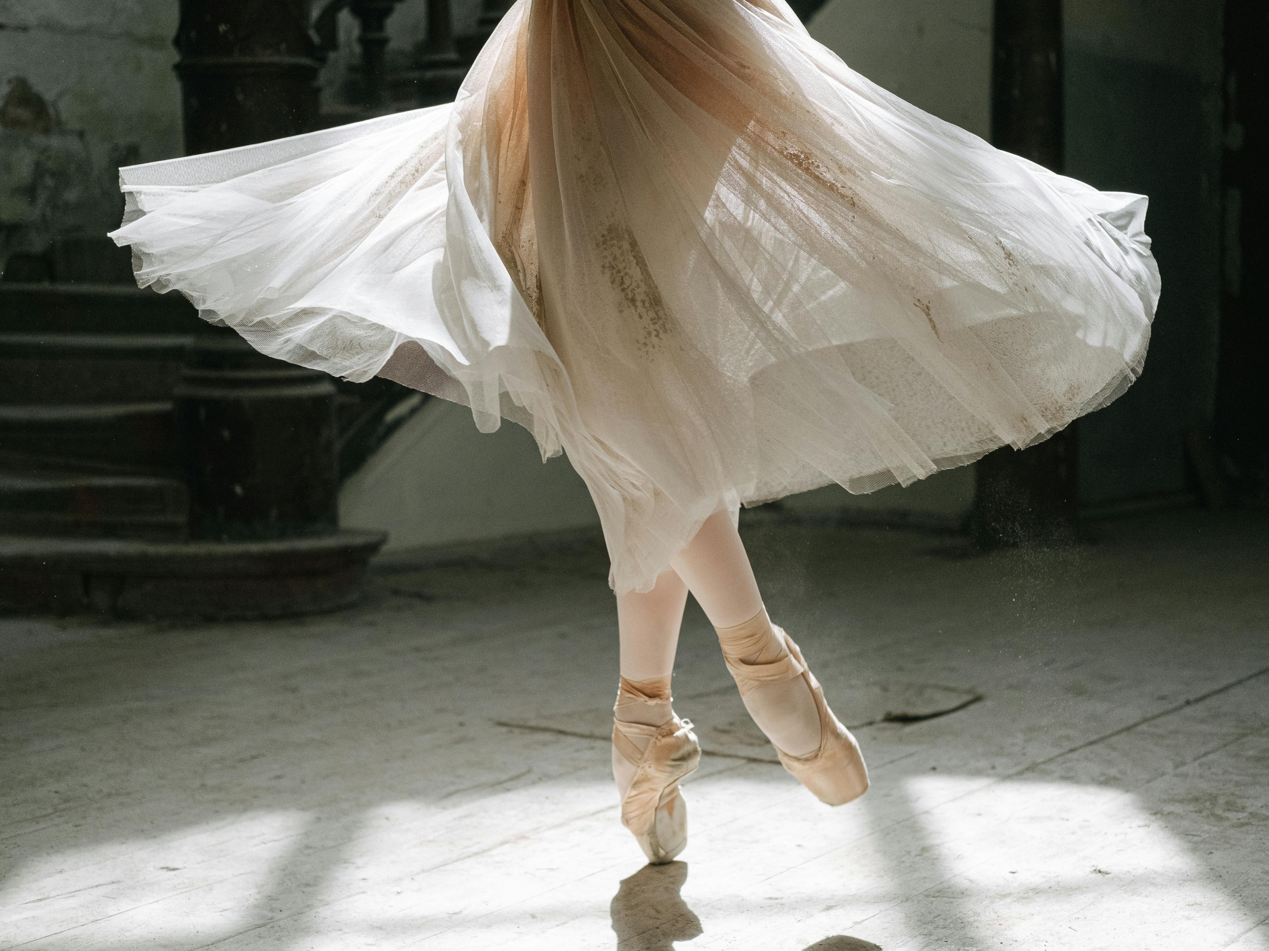 Close-up of a ballet dancer's legs and feet in pointe shoes, mid-spin, with a flowing tutu. The scene appears to be in a dance studio or theater, with a dark background.