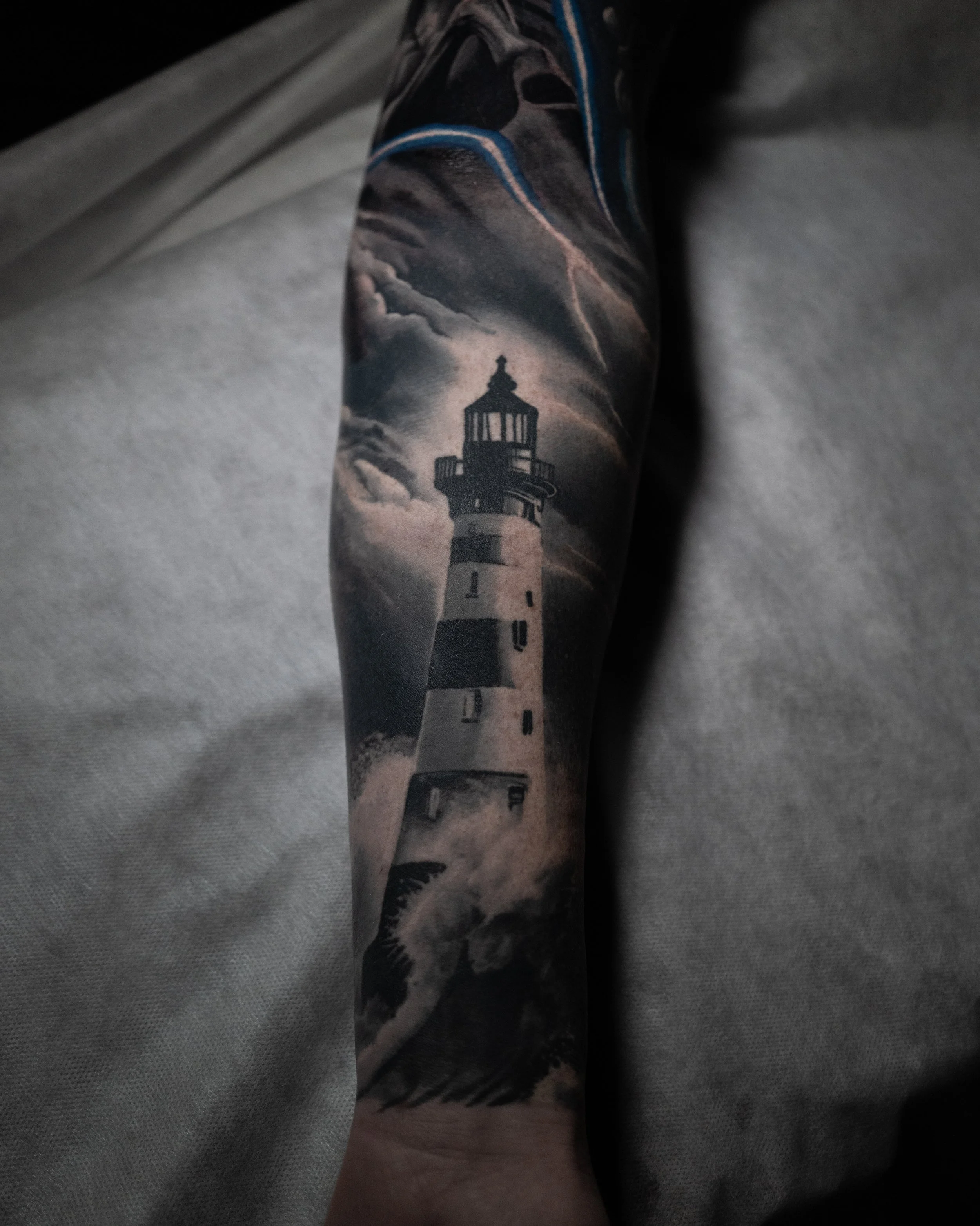 Black and white tattoo of a lighthouse on a person's arm, with clouds in the sky and waves at the base.