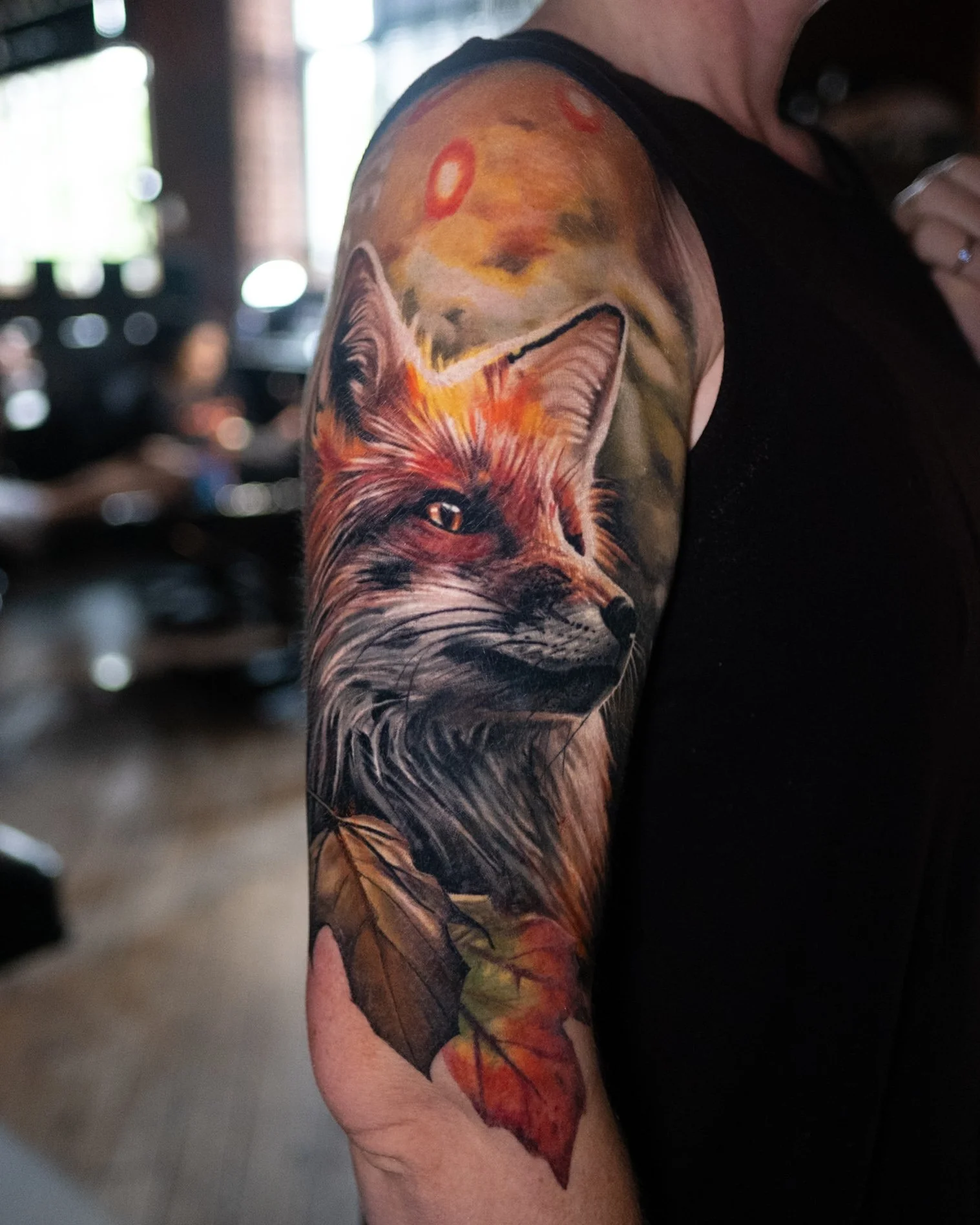 Colorful tattoo of a fox's head with a background of sunset and autumn leaves on a person's upper arm.