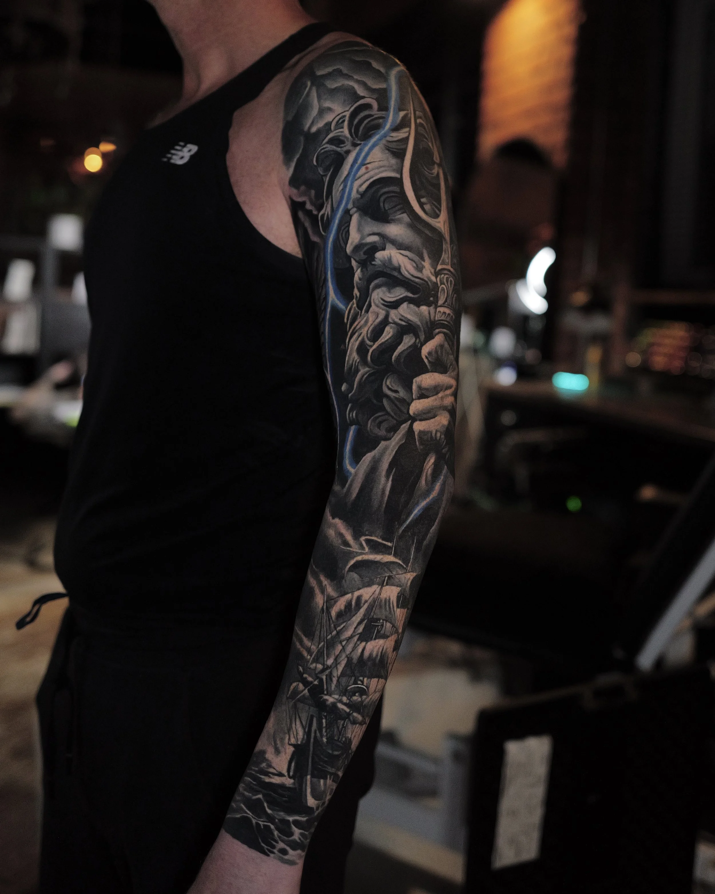 Tattoo of an old man with a beard, holding a staff, with a stormy sky above. Below, a portrait of a sailing ship at sea with waves.