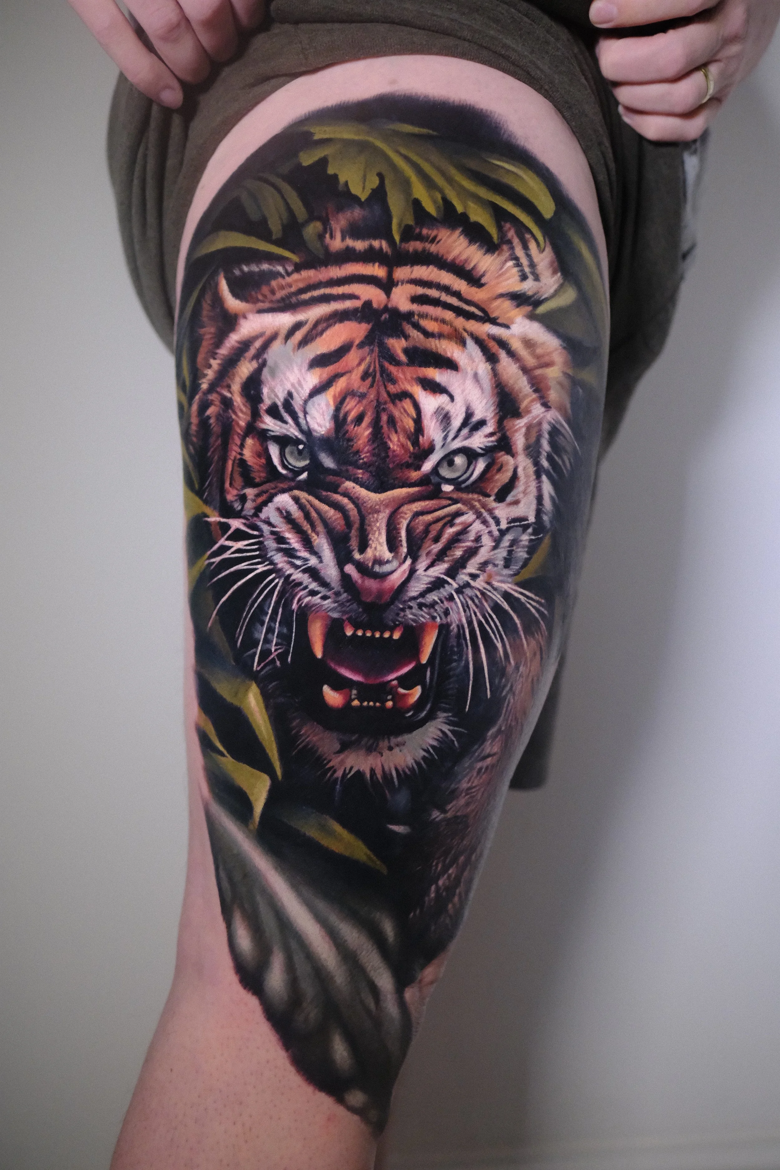 Tattoo of a fierce tiger's face on person's thigh, with green leaves in the background.